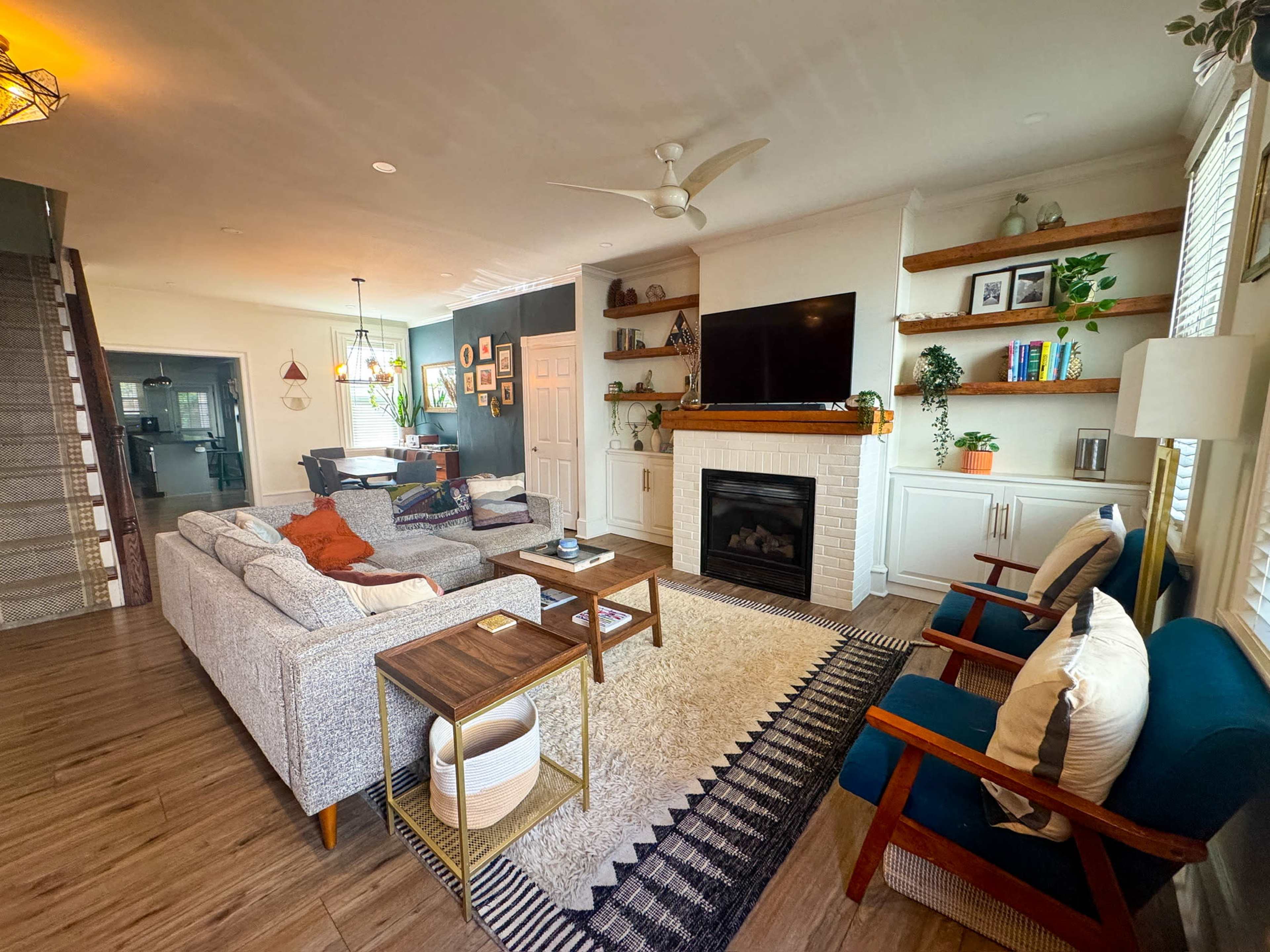 The image shows a modern living room with a fireplace, a large television mounted on a wall, and various seating arrangements, including sofas and chairs surrounded by wooden shelves and decor items.
