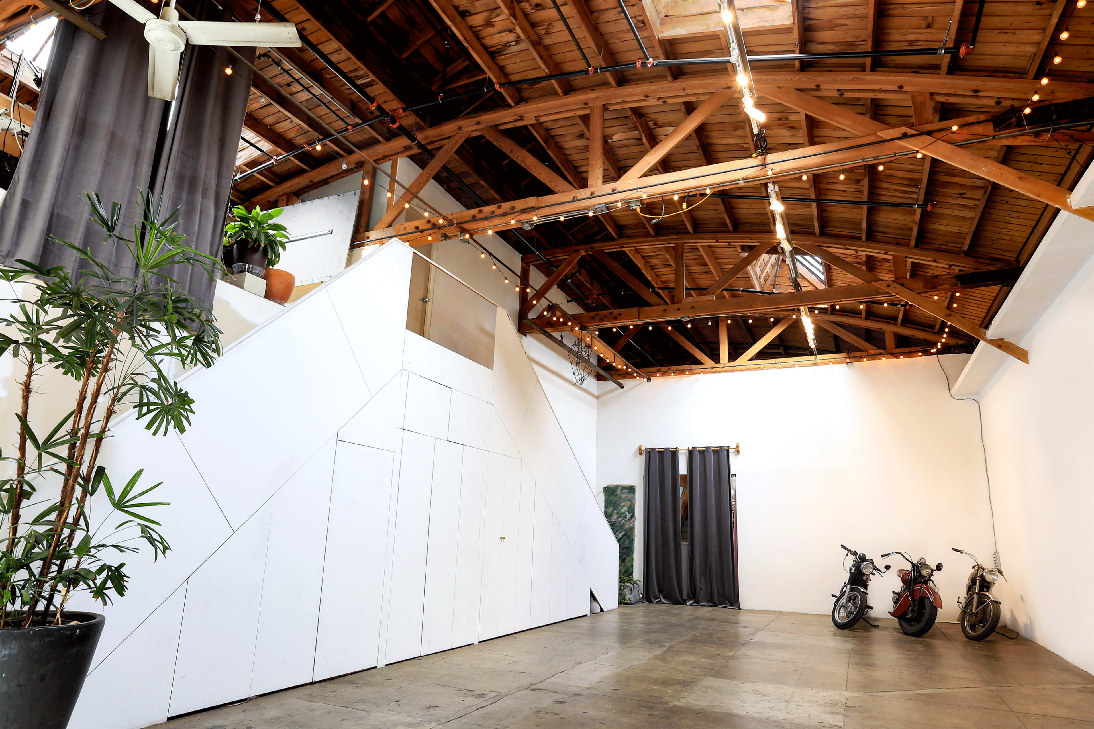 The image shows an interior space with a high wooden ceiling, large windows, and two parked bicycles near a wall, along with decorative plants and draped curtains.