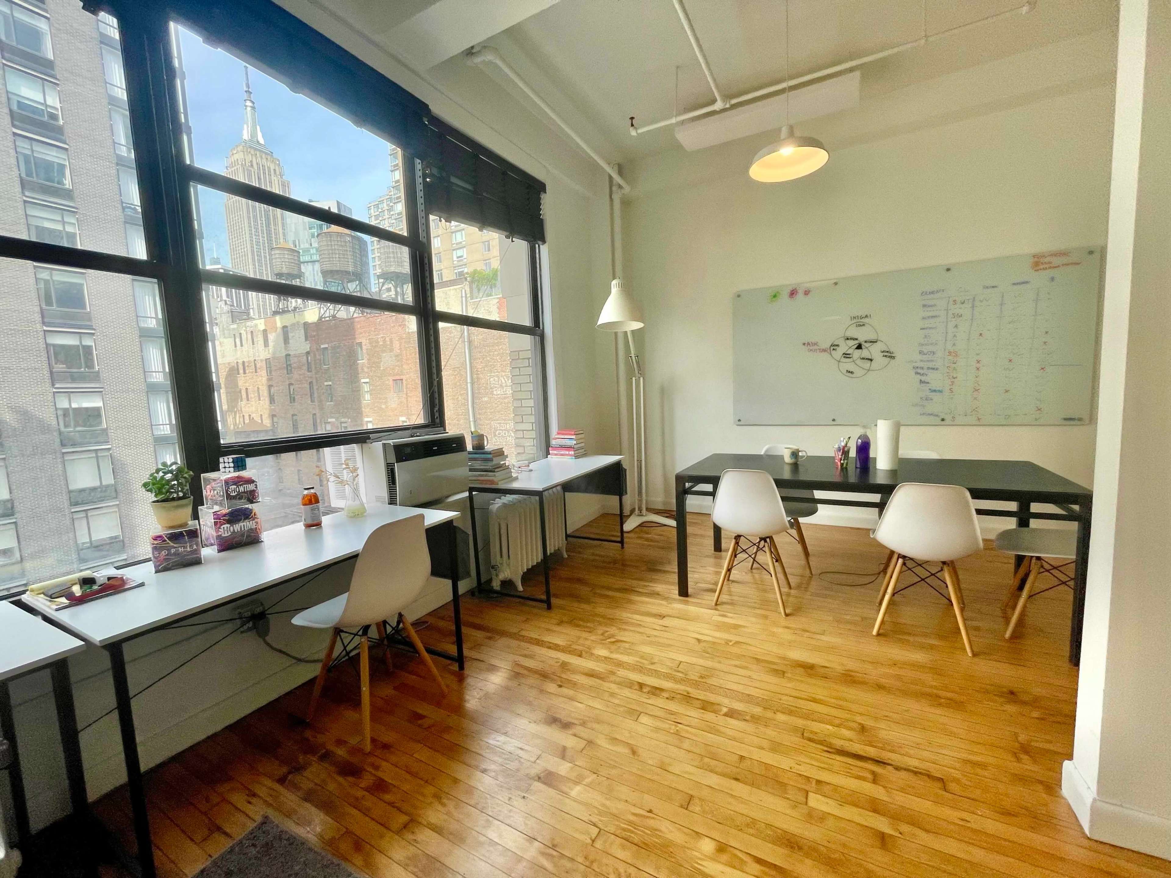 A bright office features large windows with a view of the Empire State Building, hardwood floors, two workstations, and a whiteboard on the wall.