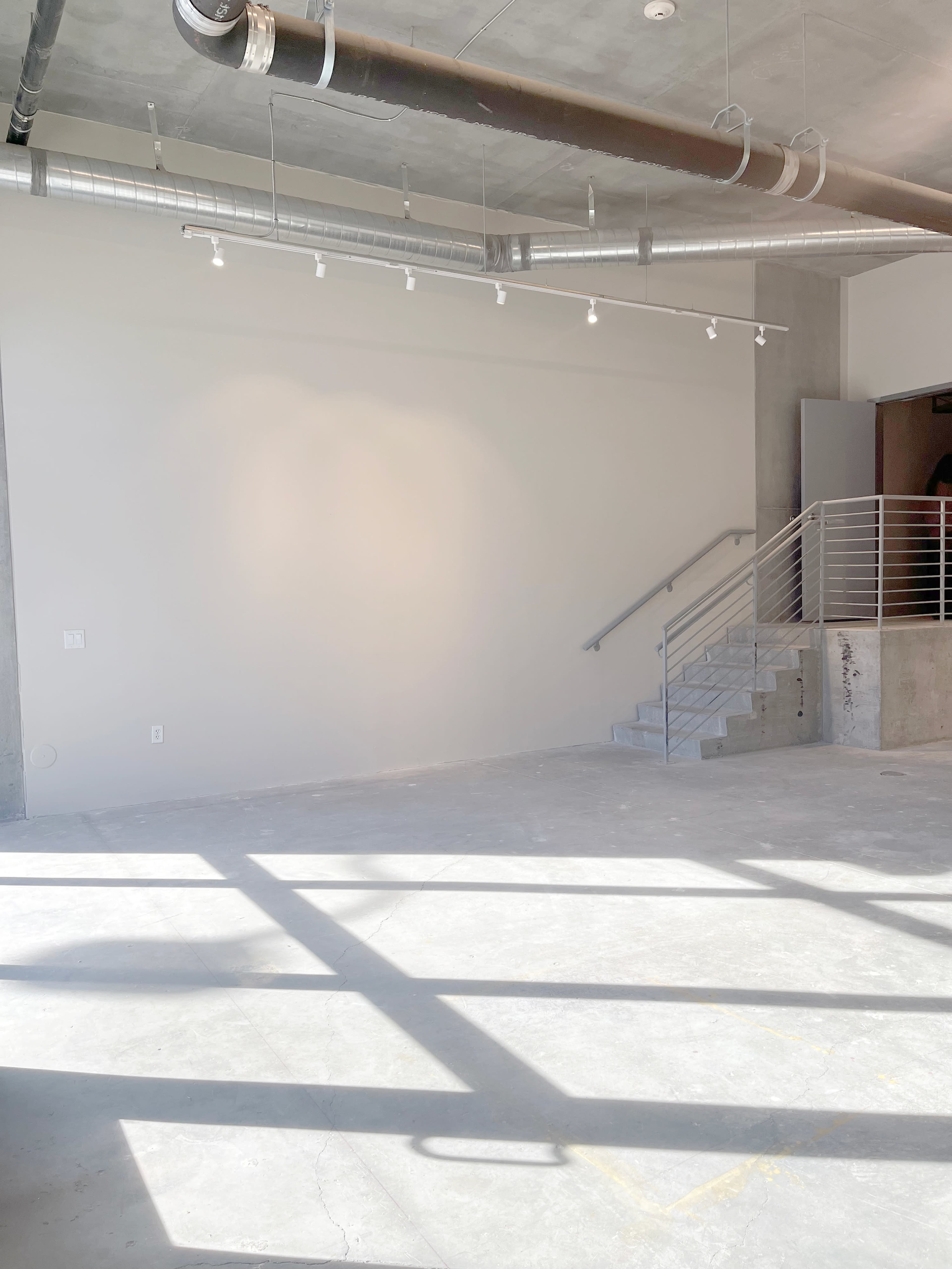 The image shows a vacant, industrial-style room with concrete flooring, a blank wall, and a staircase leading to an upper level.