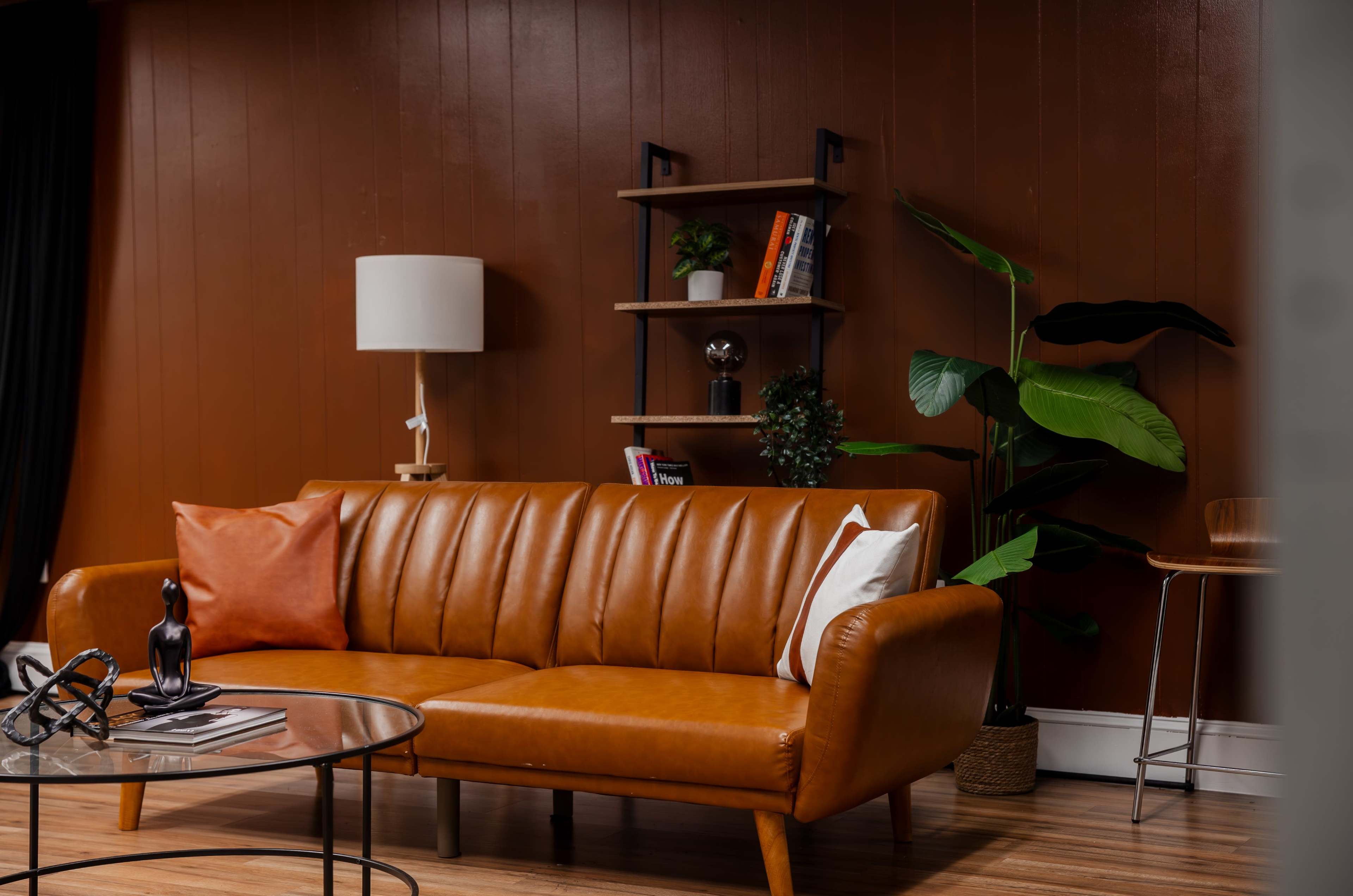 A brown leather couch with decorative pillows is positioned in a room alongside a wooden bookshelf, a standing lamp, and a large indoor plant.