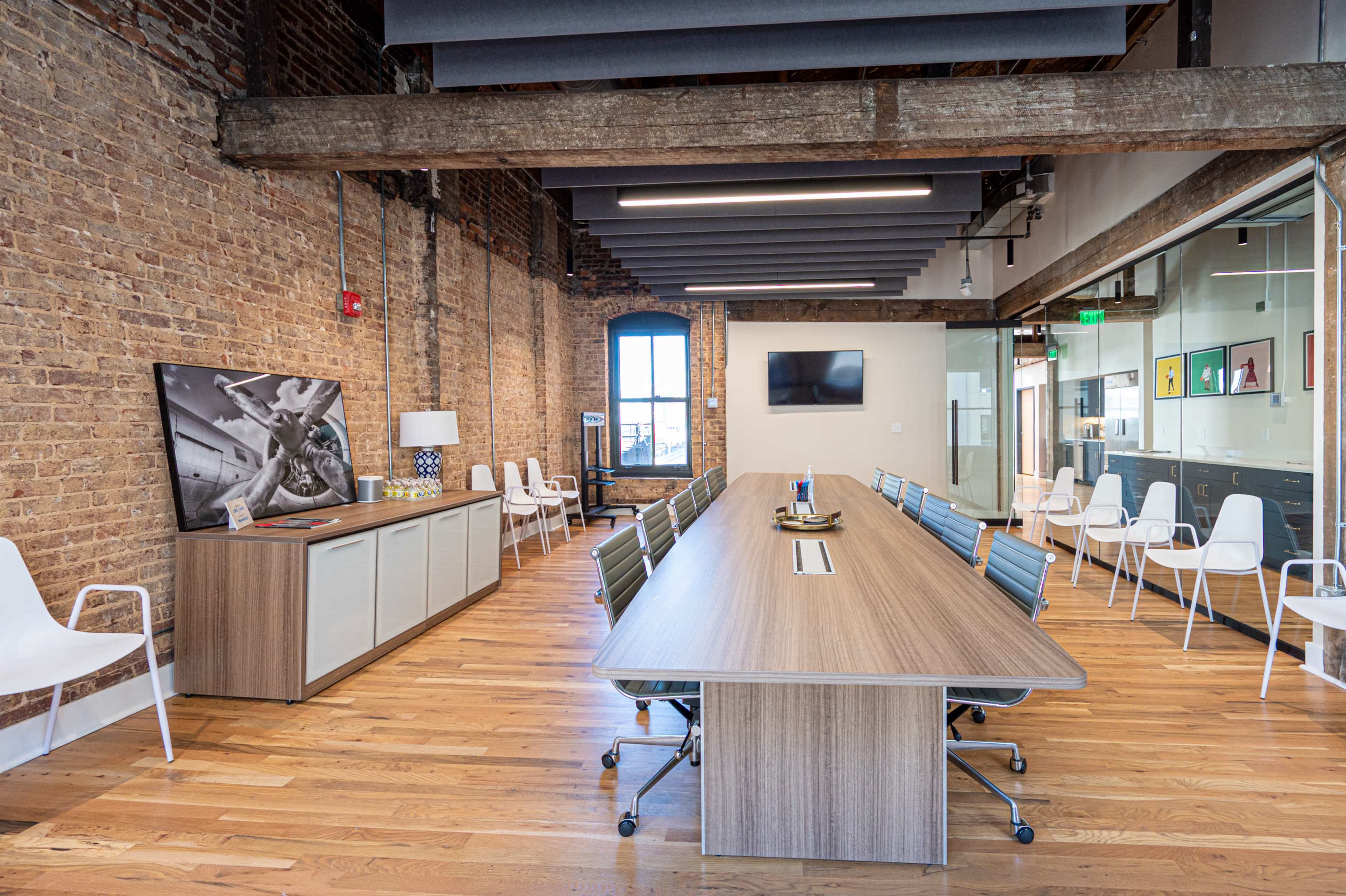 A modern conference room with a long wooden table, white chairs, exposed brick walls, and large windows.
