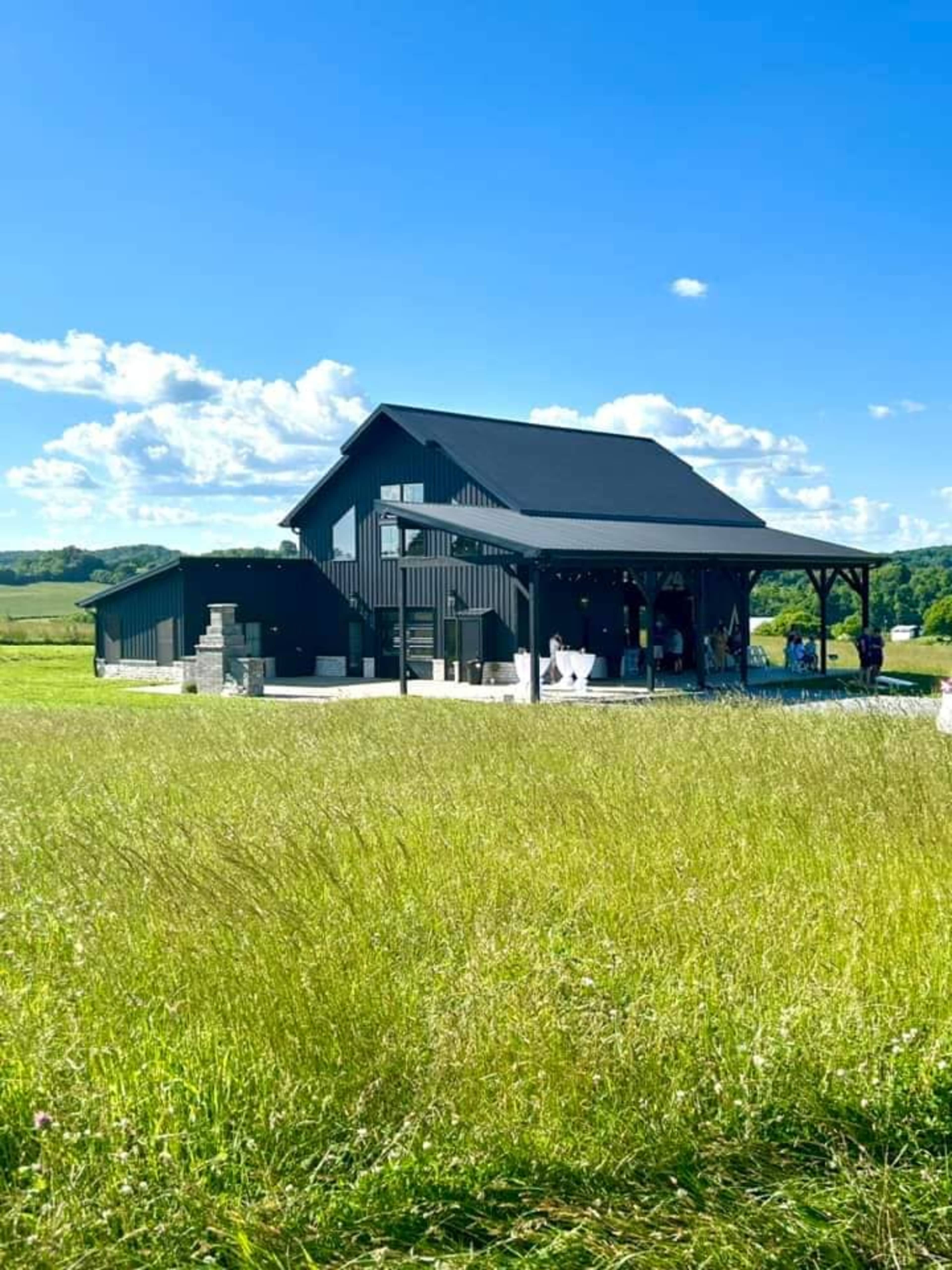 A large black barn-style structure with a covered porch sits amidst a green field under a bright blue sky.