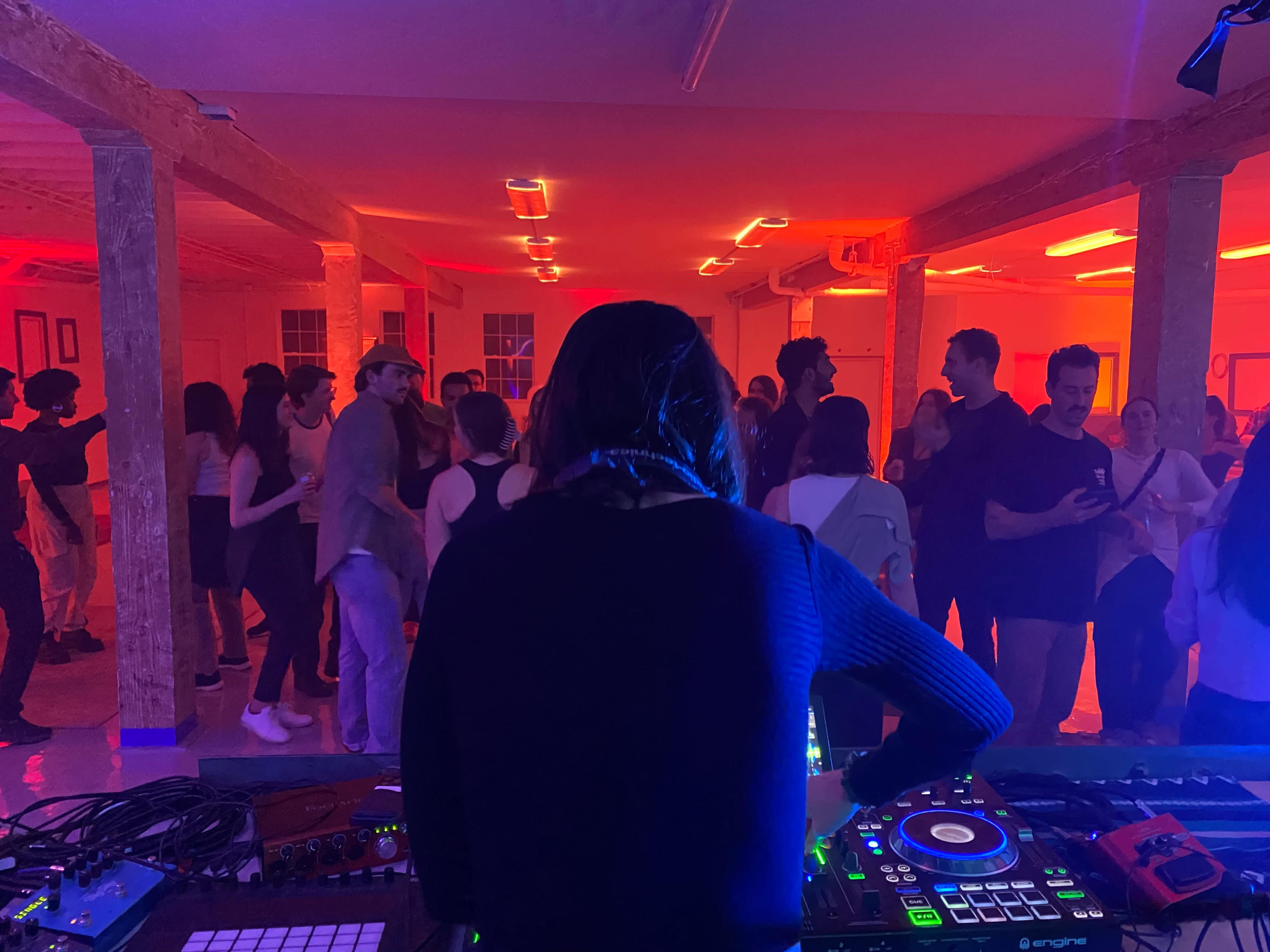 A DJ stands at a mixing console while a crowd dances in a dimly lit room with colorful lighting.