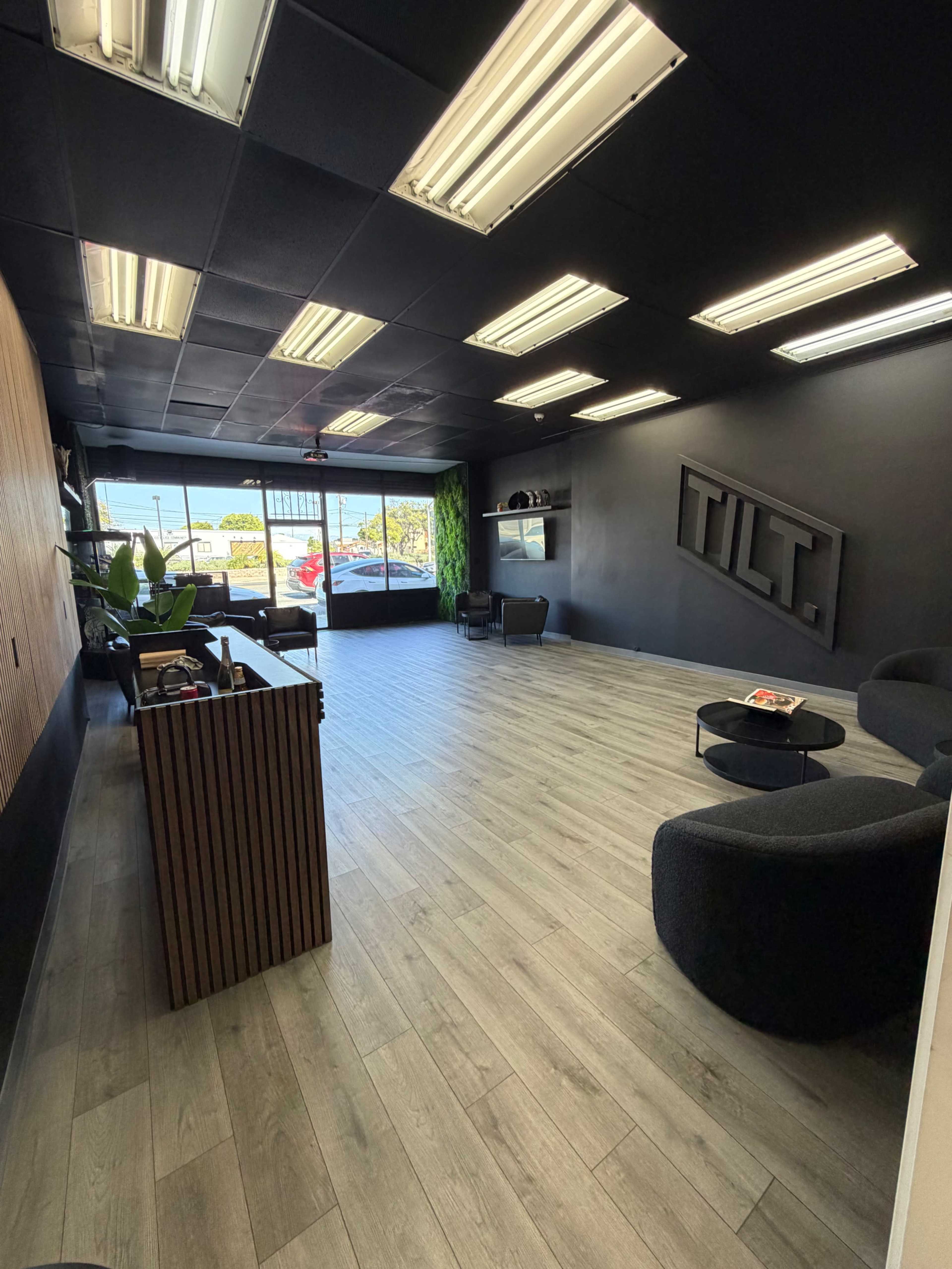 The image shows a modern office reception area with dark walls, wooden paneling, and minimalist seating arrangements near a large glass entrance.