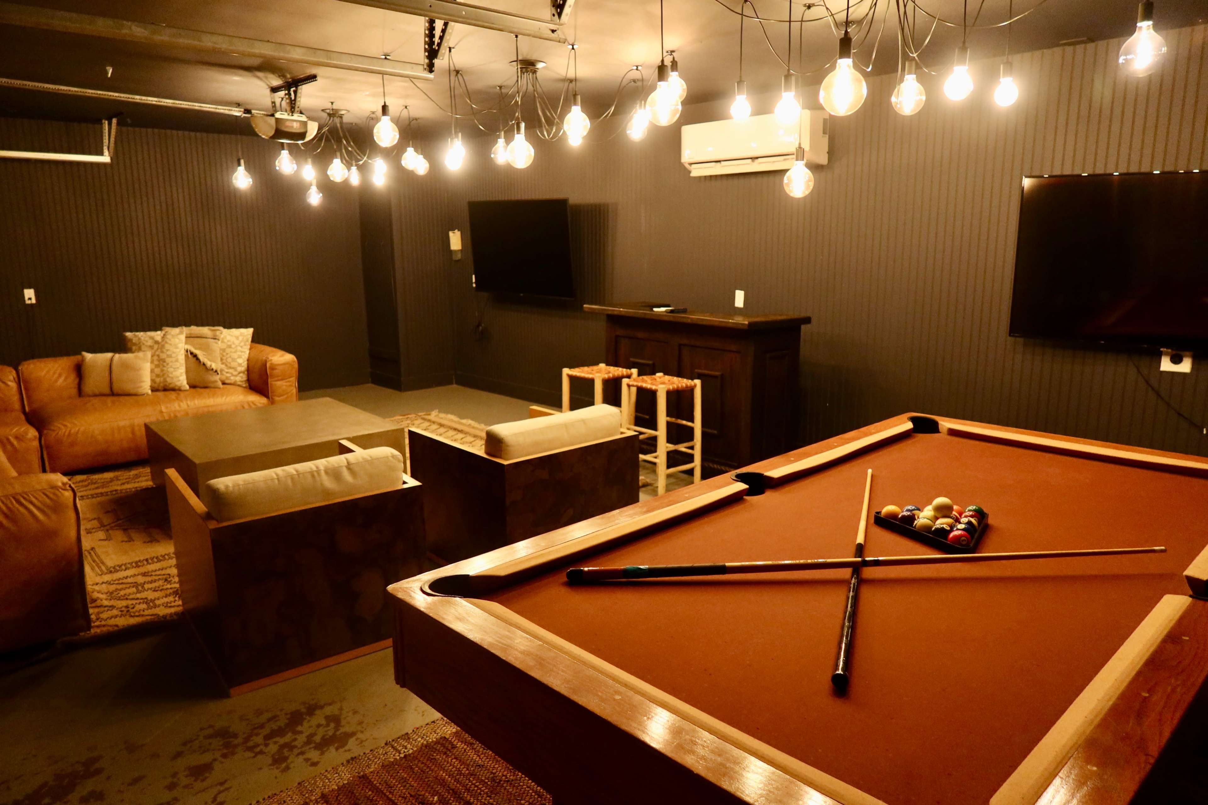 The image shows a game room featuring a pool table in the foreground, a comfortable seating area, and multiple light fixtures hanging from the ceiling.