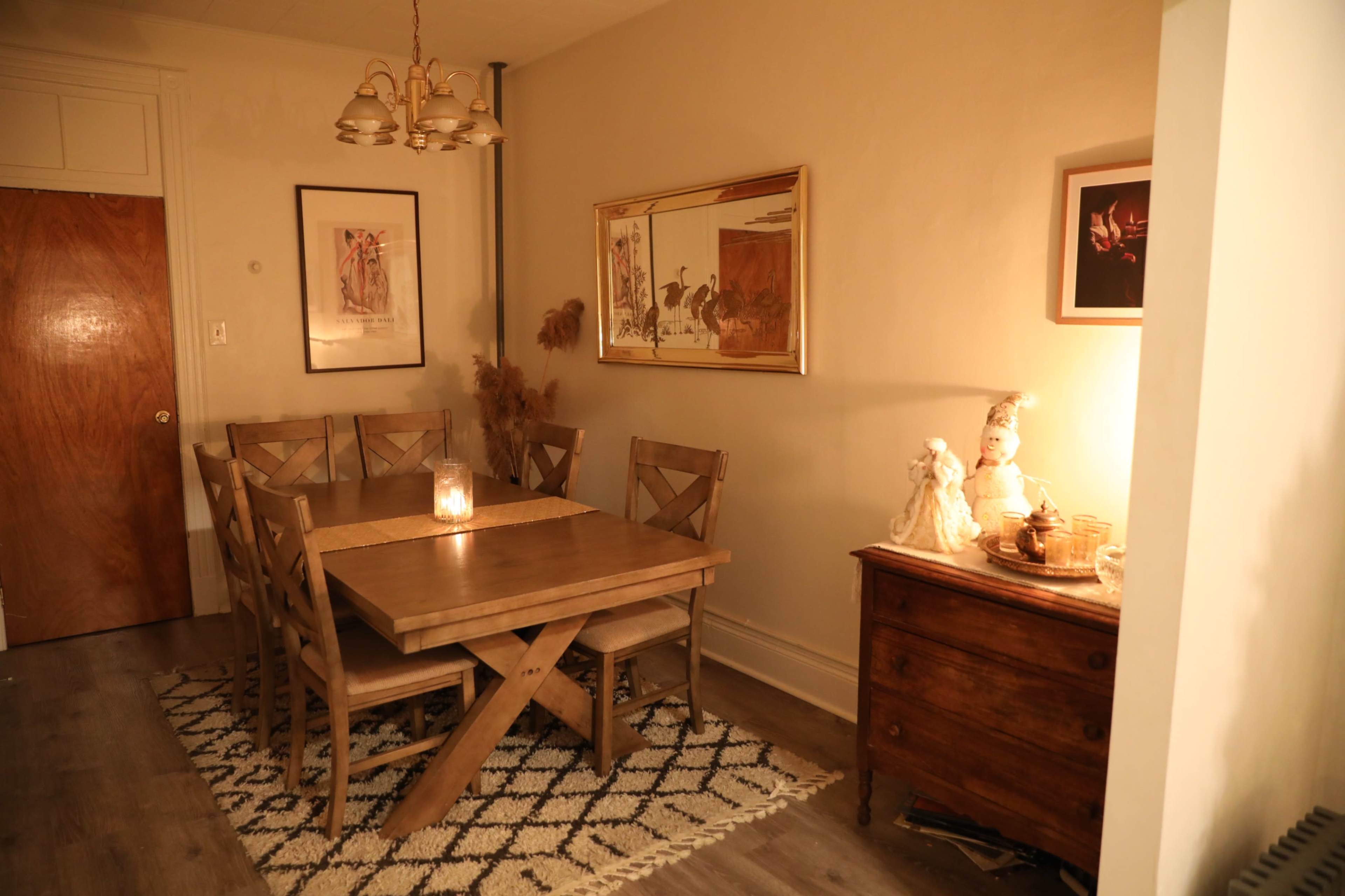 The image depicts a warmly lit dining area featuring a wooden table surrounded by chairs, with artwork on the walls and a decorative side table.