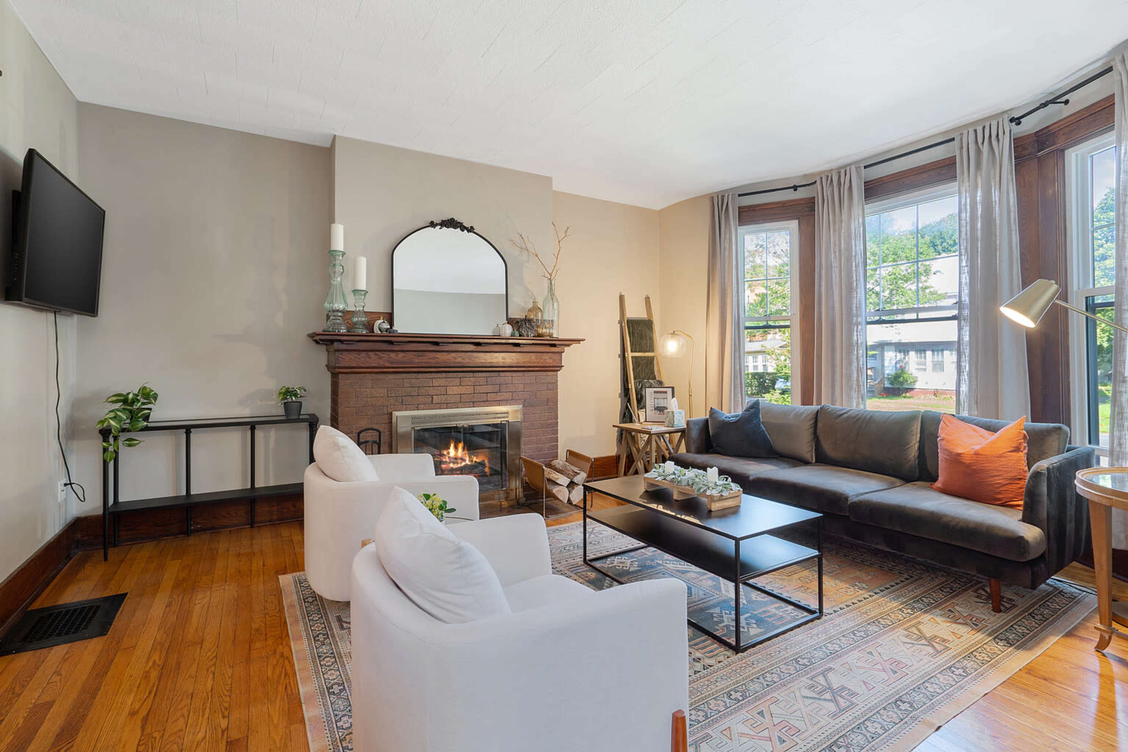 The living room features a cozy seating arrangement with a dark sofa and two white armchairs, a brick fireplace, and large windows allowing natural light to enter.