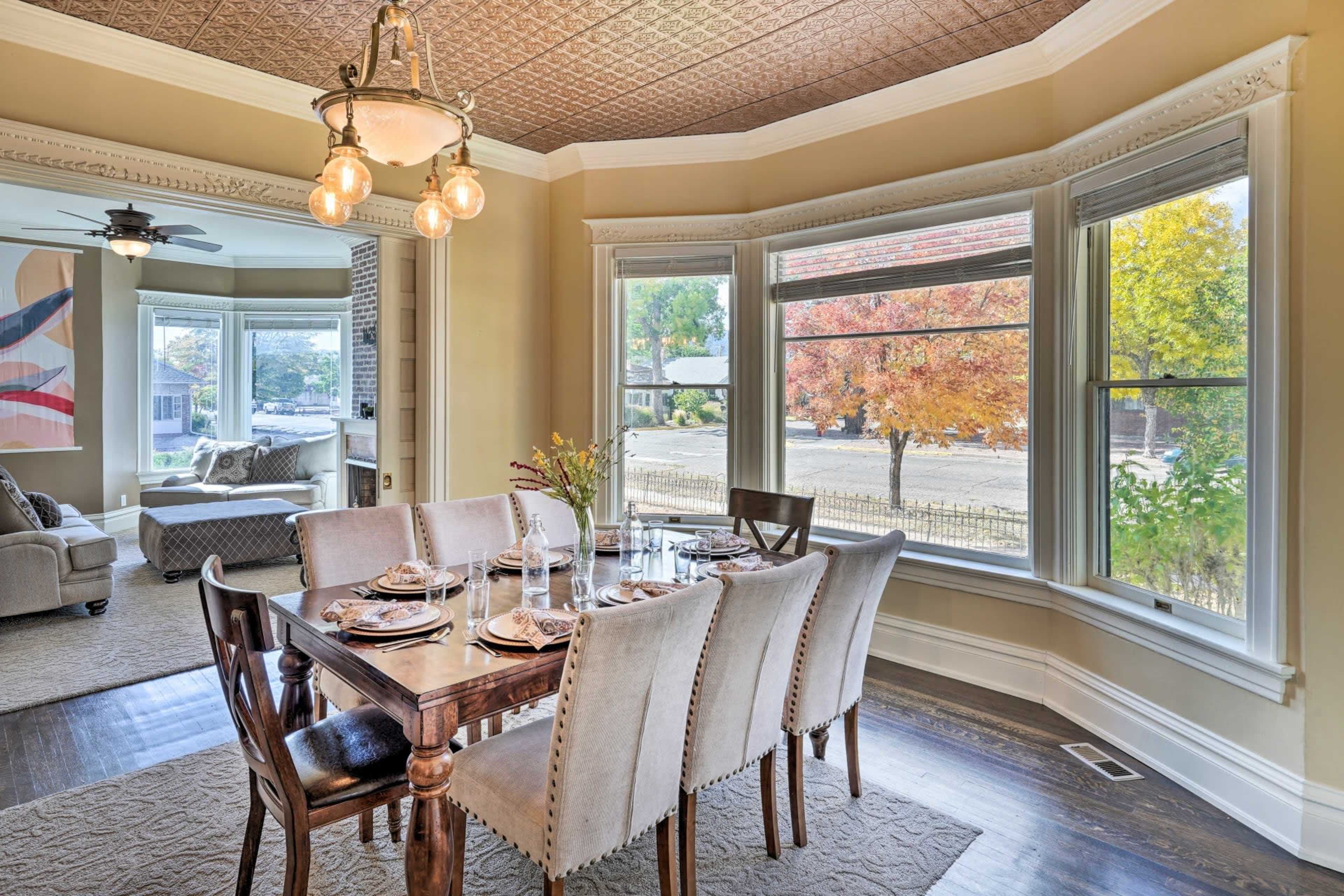 A dining room with a wooden table set for a meal, surrounded by upholstered chairs, features large bay windows that overlook a tree-lined street.