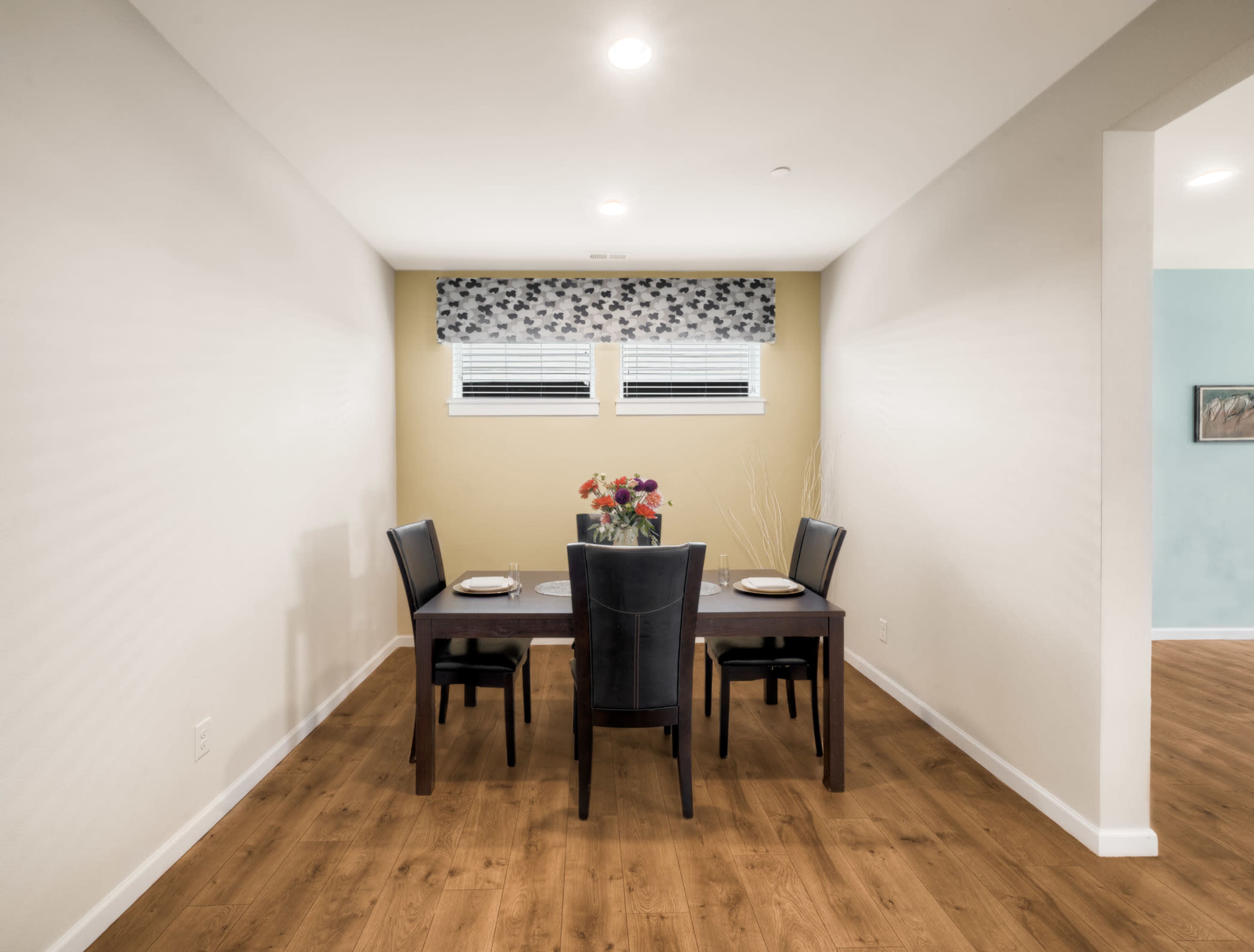 A dining area features a dark wooden table set for four, with black chairs and a floral centerpiece, illuminated by ceiling lights and decorated with simple window treatments.