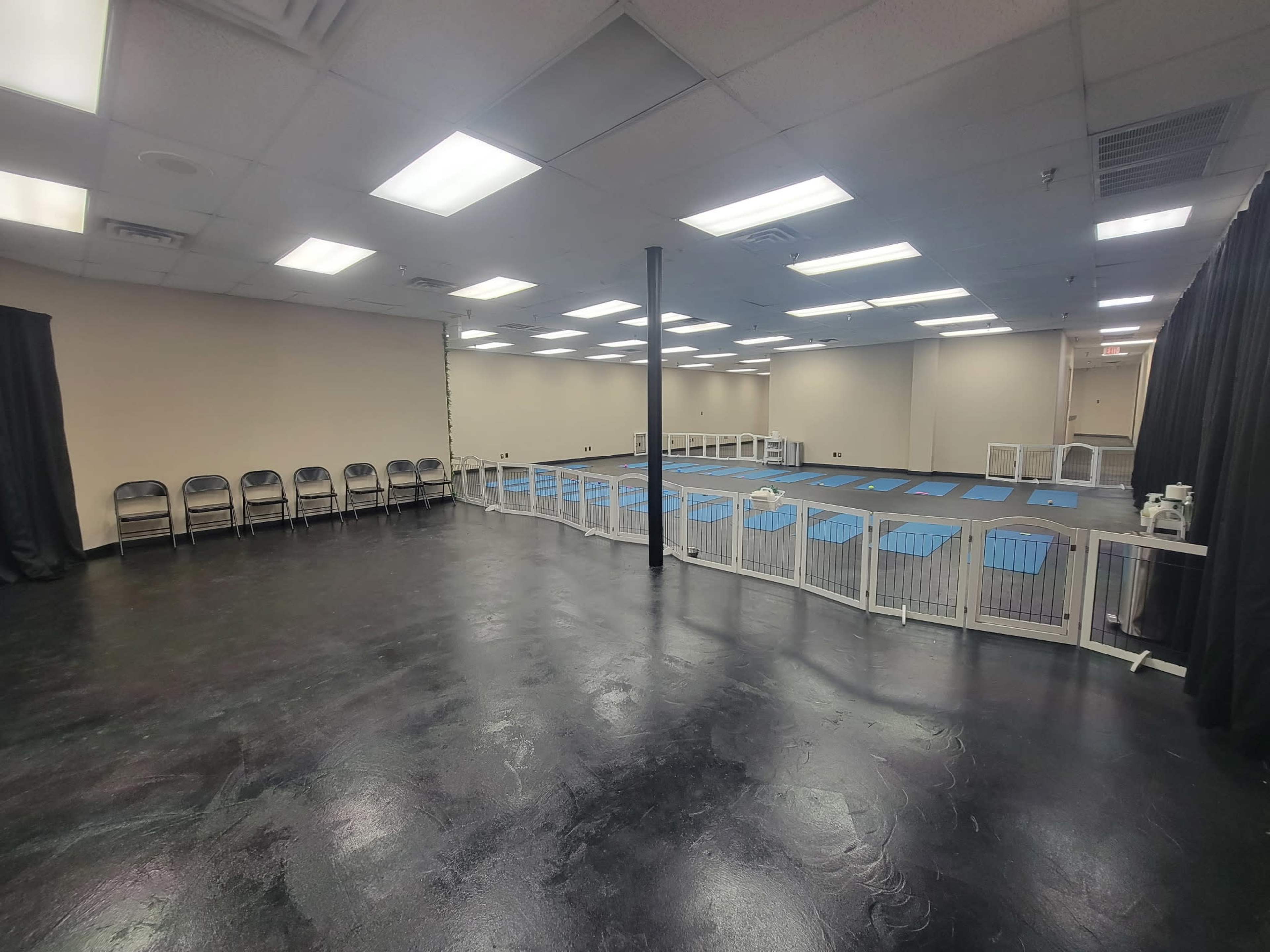 The image shows a spacious room with a polished floor, lined with folding chairs on one side and a fenced area with blue mats in the center.