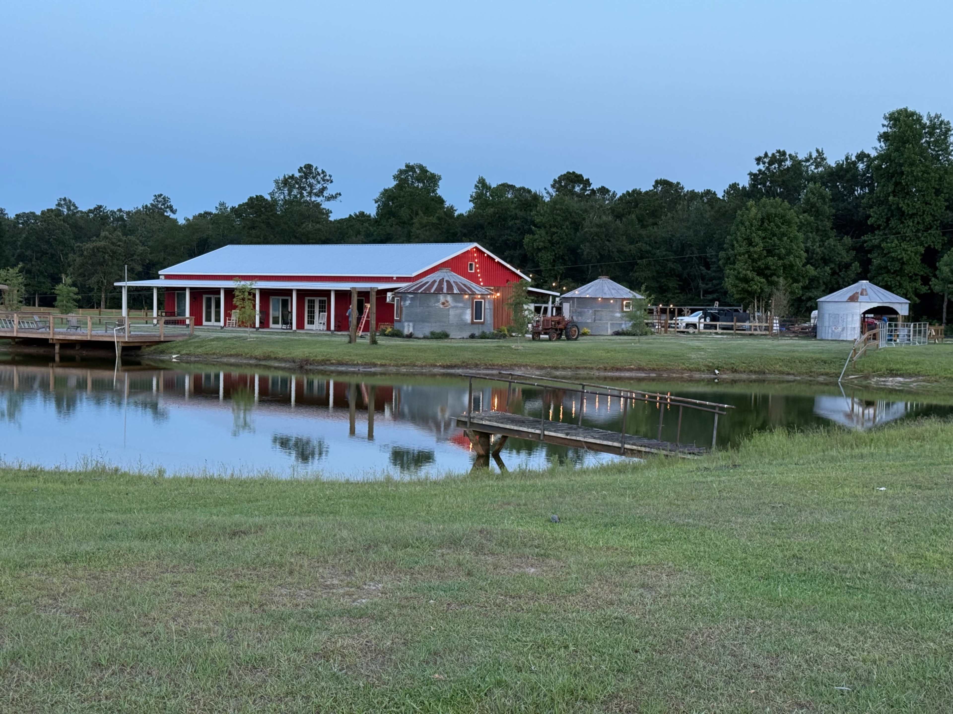 A red building with a white roof stands near a pond, reflecting in the water, with additional structures and trees in the background.