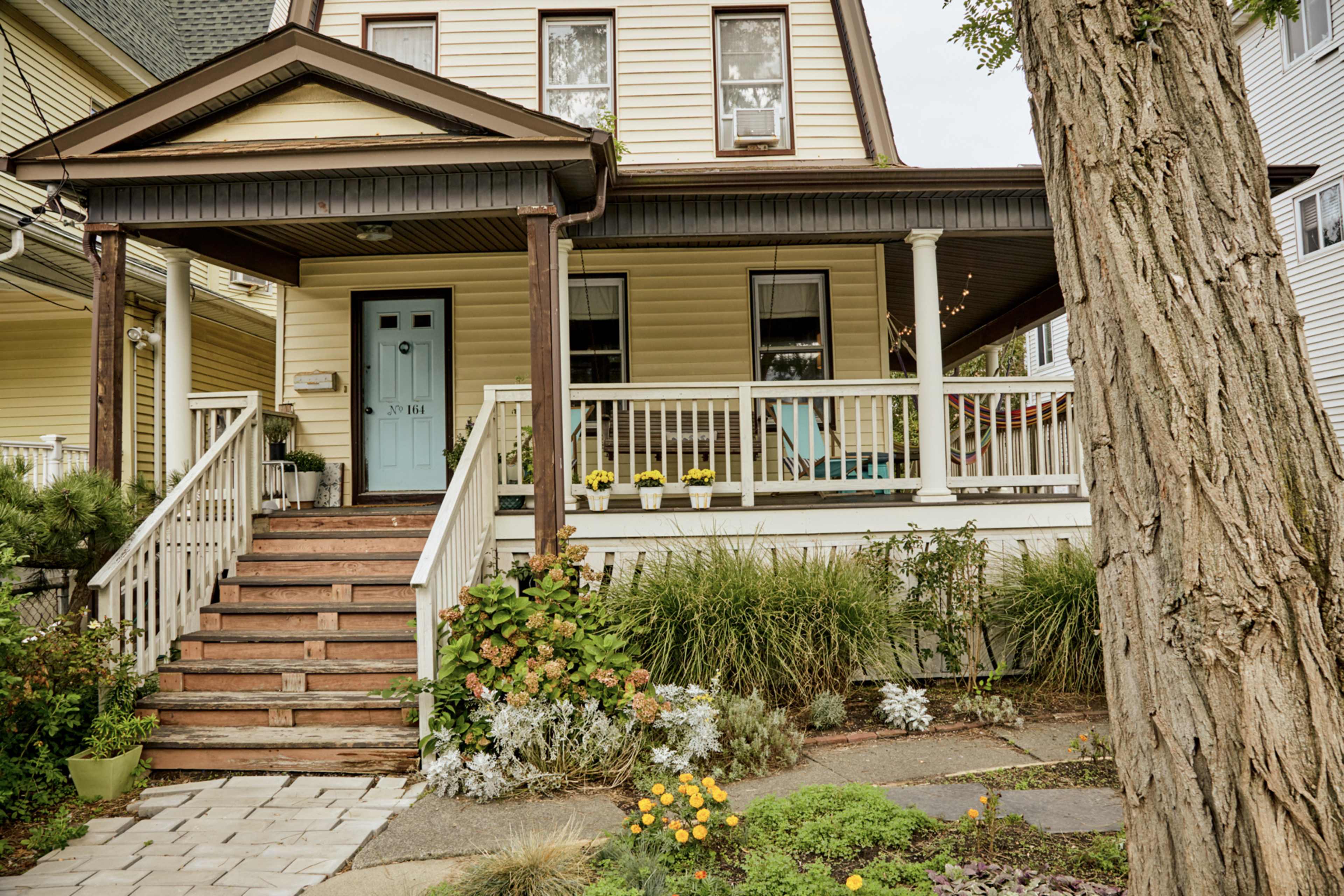 A two-story house with a front porch, wooden stairs, a blue door, and a garden filled with flowering plants.