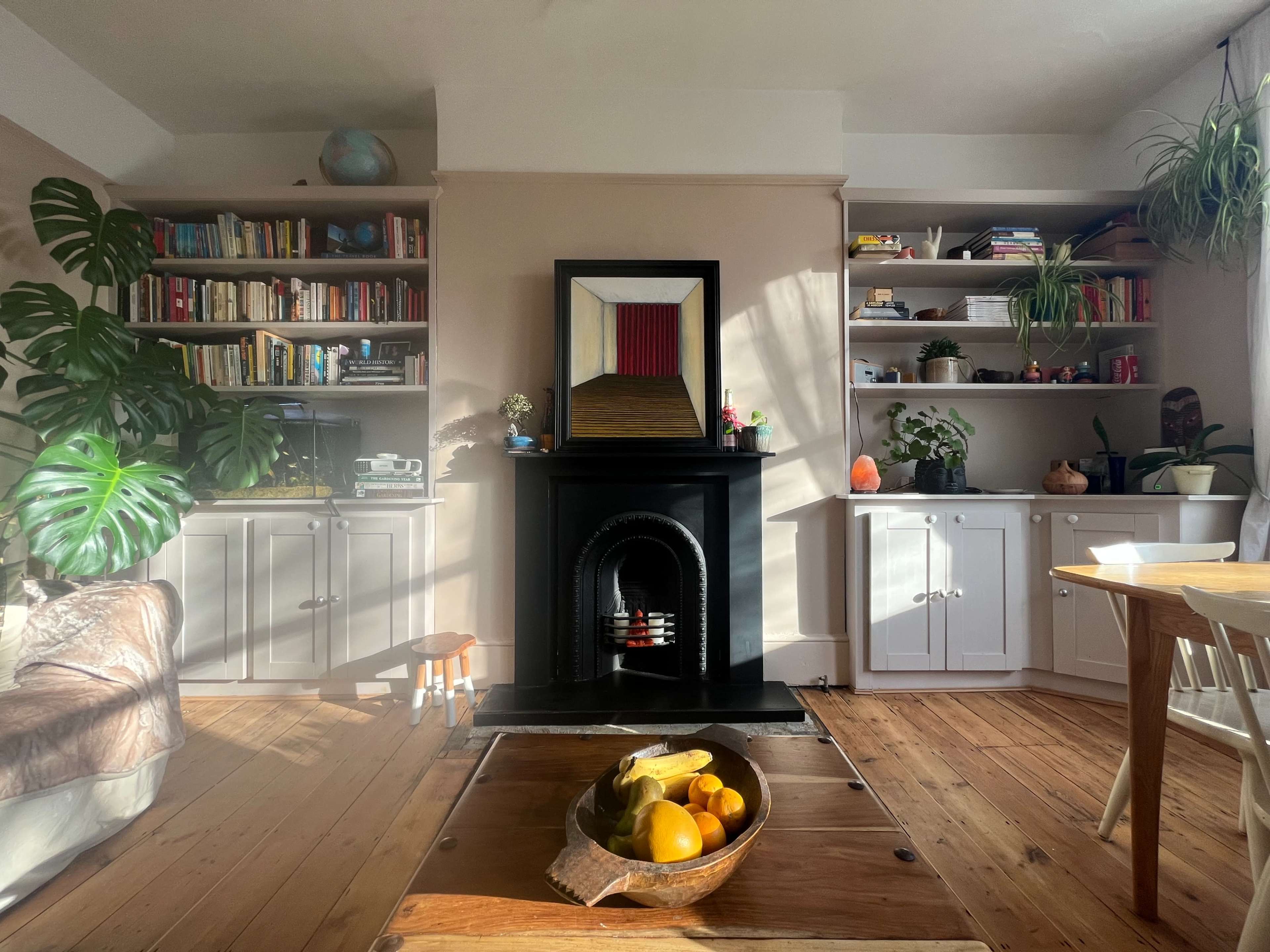 A cozy living room features a black fireplace, a large painting, bookshelves filled with books, and a dining table with a bowl of fruit.