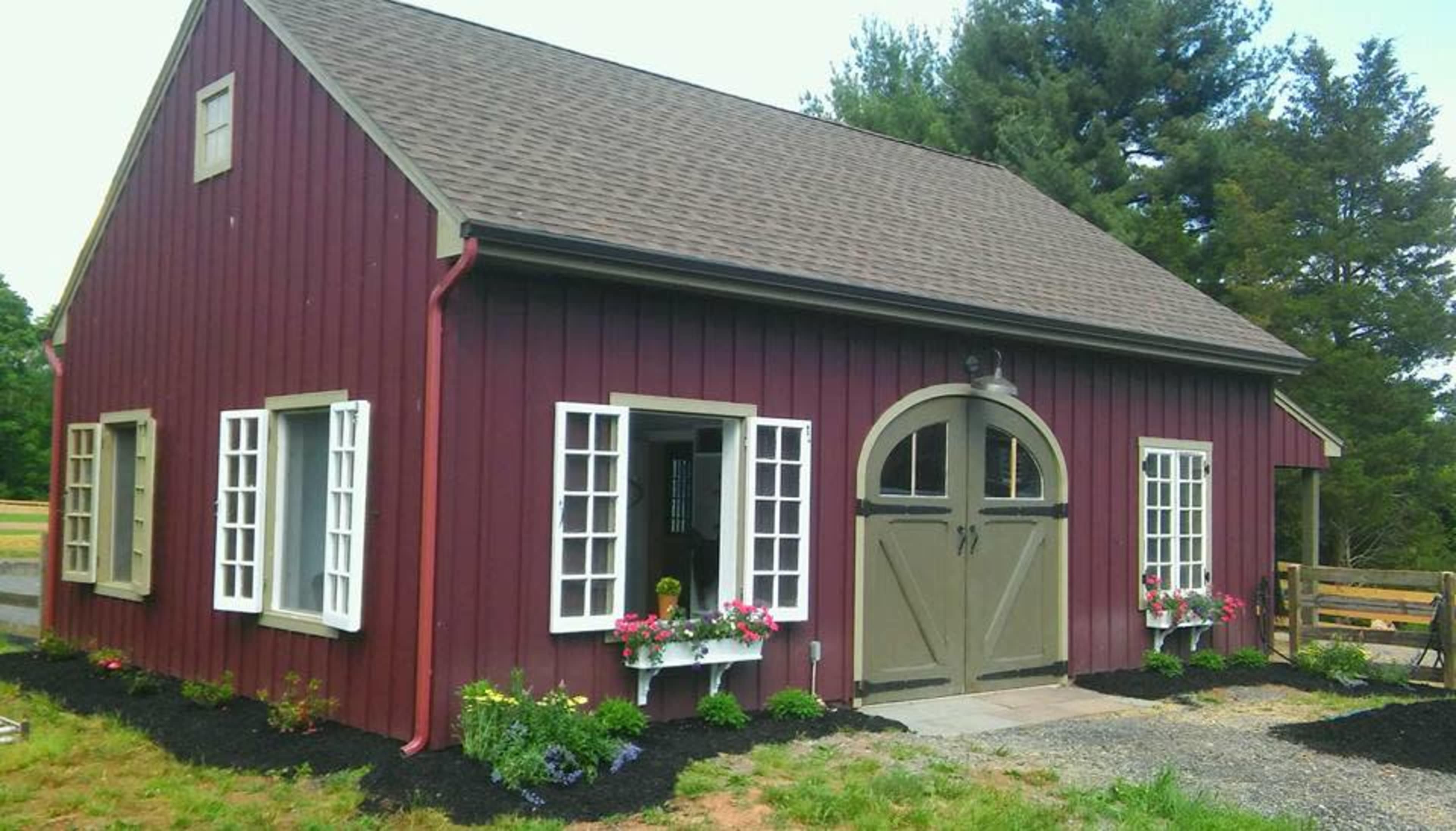 A red wooden house with a steep roof features large arched double doors and multiple windows, surrounded by flower boxes and gravel landscaping.