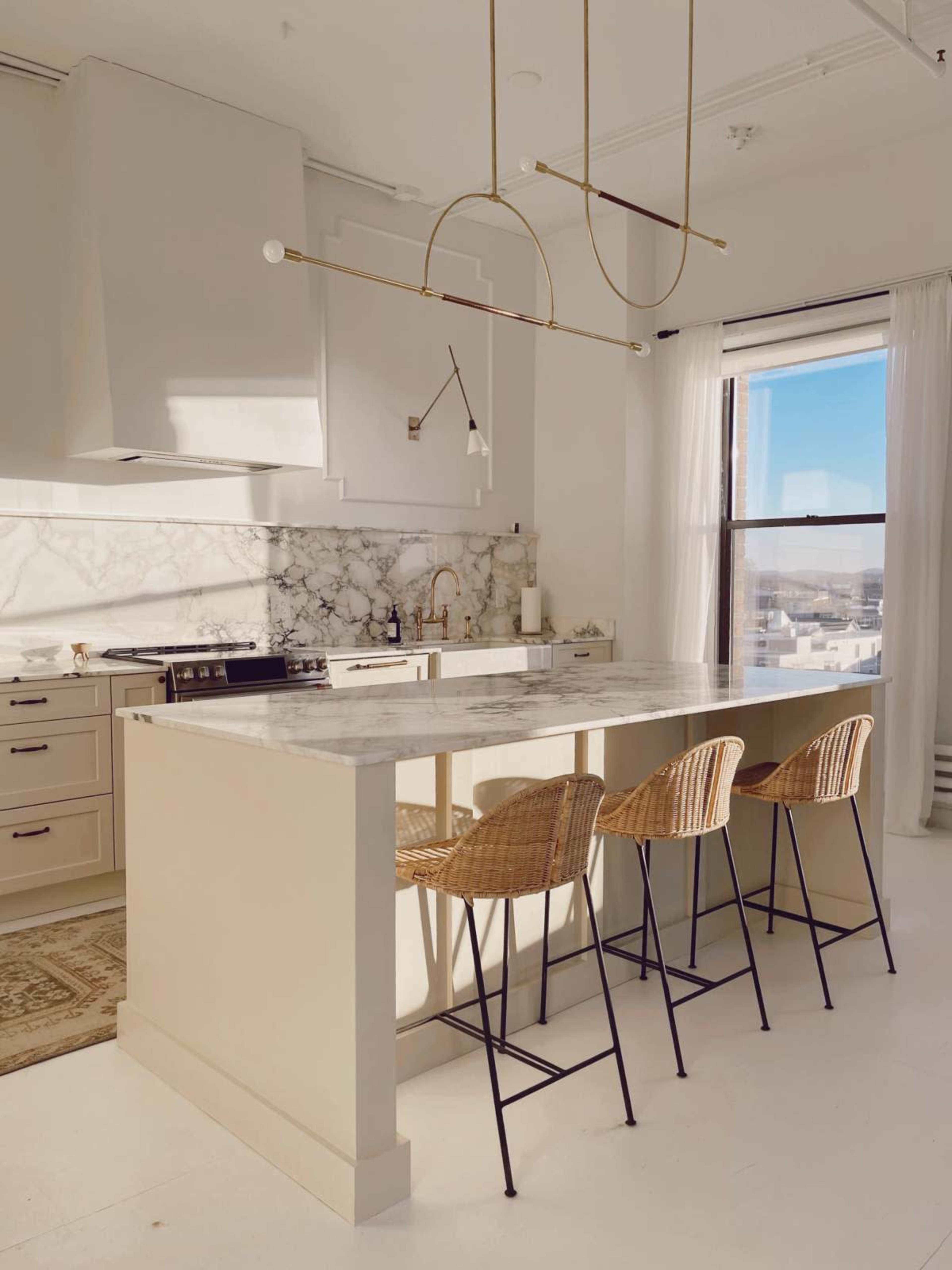 The image shows a modern kitchen with a large marble-topped island and three metal and wicker bar stools.