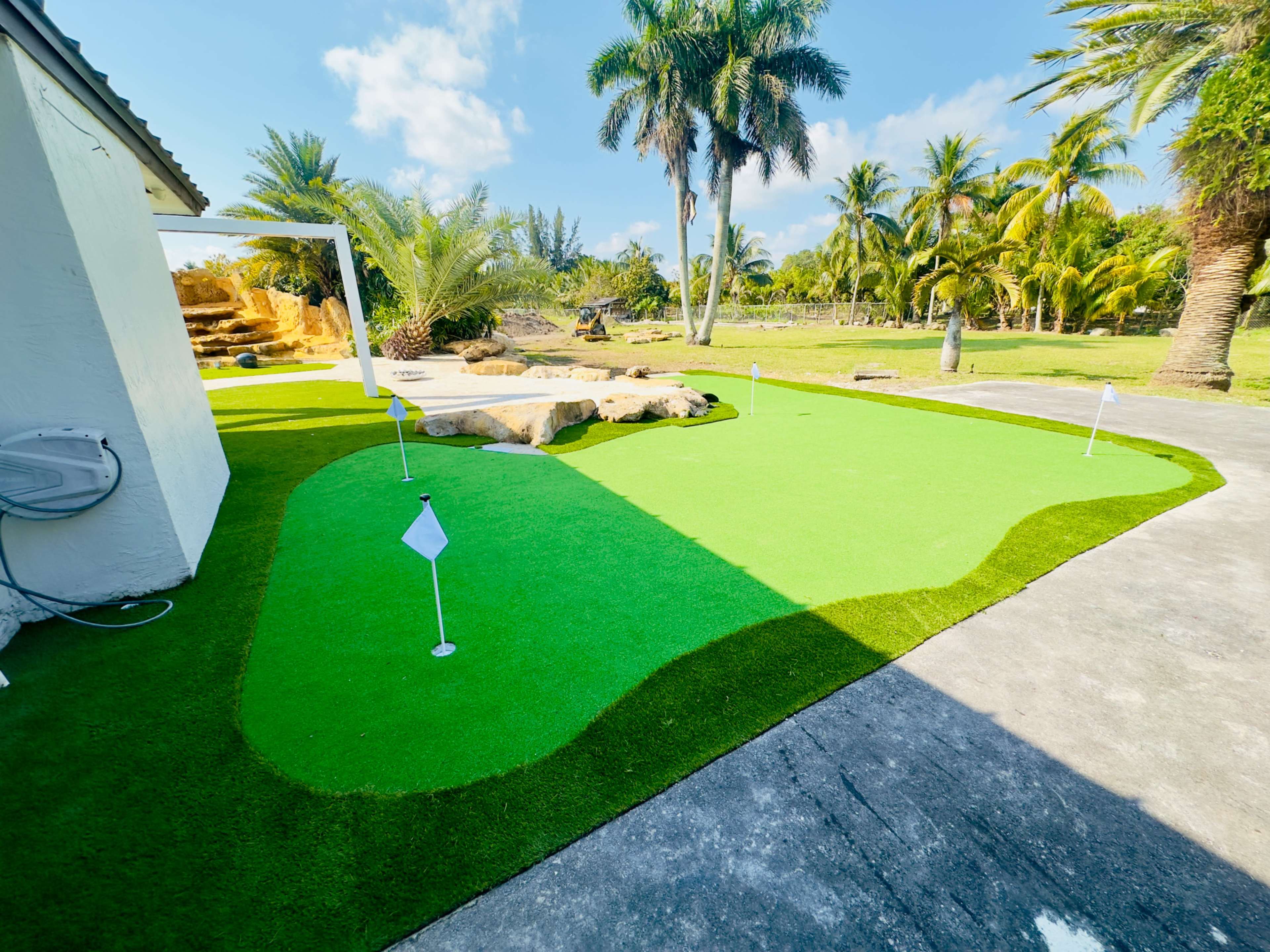 The image shows a small putting green with white flags on a well-maintained artificial grass surface, surrounded by palm trees and a clear blue sky.