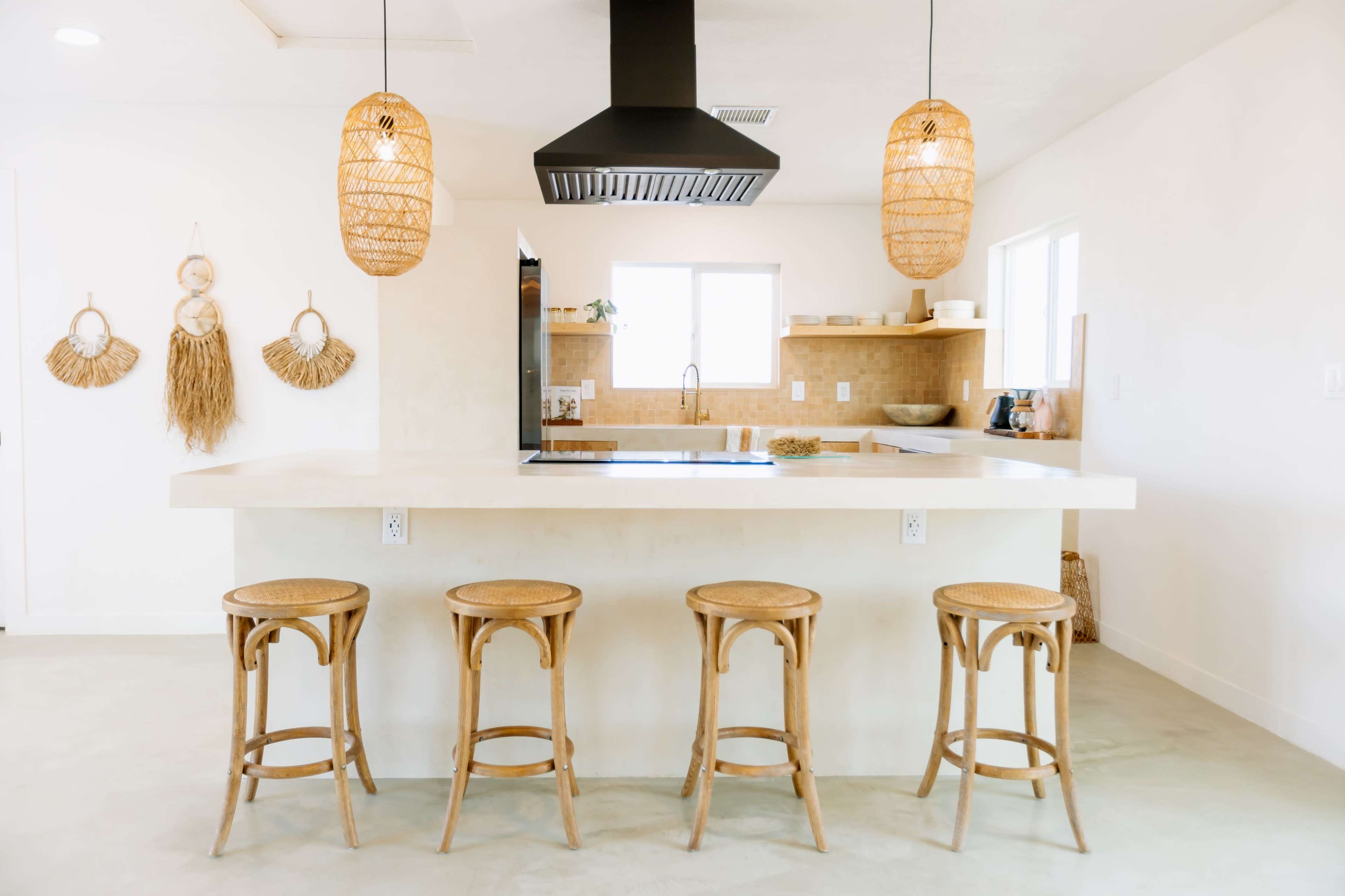 A modern kitchen features a light-colored island with four woven bar stools and a black exhaust hood above the cooking area.