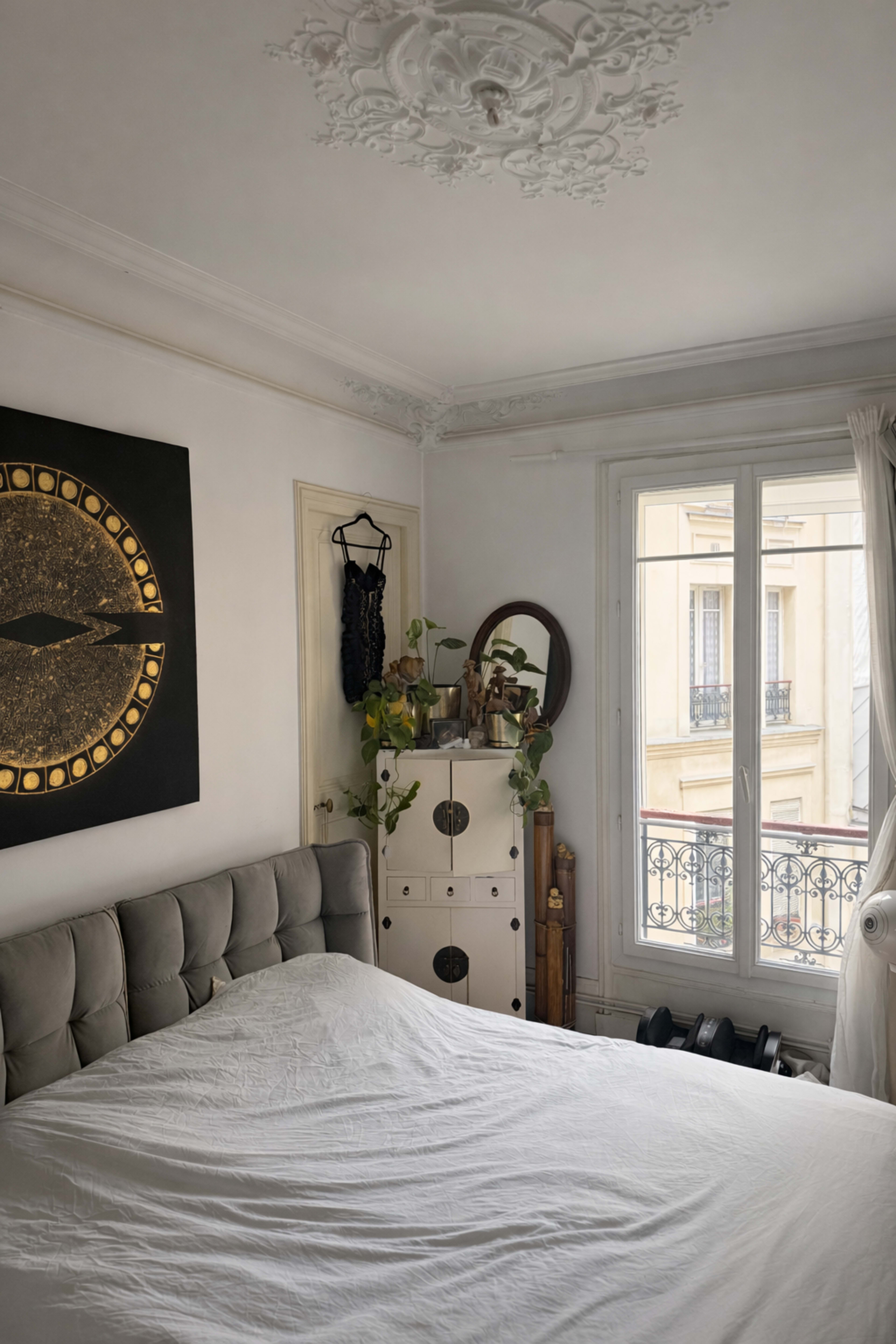 A neatly arranged bedroom features a bed with a white blanket, a decorative wall clock painting, potted plants, and a window overlooking an exterior view.