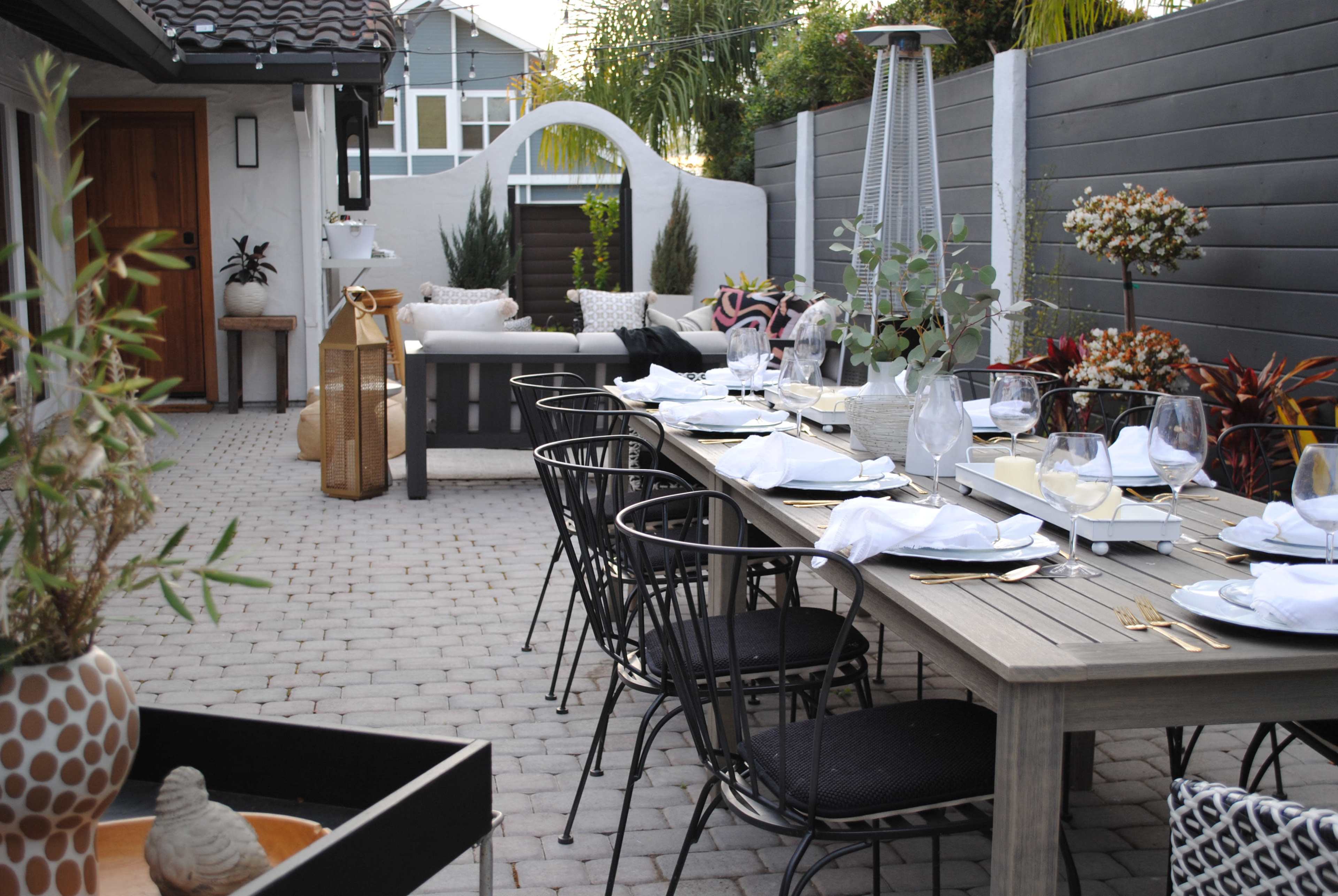 A modern outdoor dining area features a long table set with dishes and glasses, surrounded by black chairs, with a decorative garden in the background.