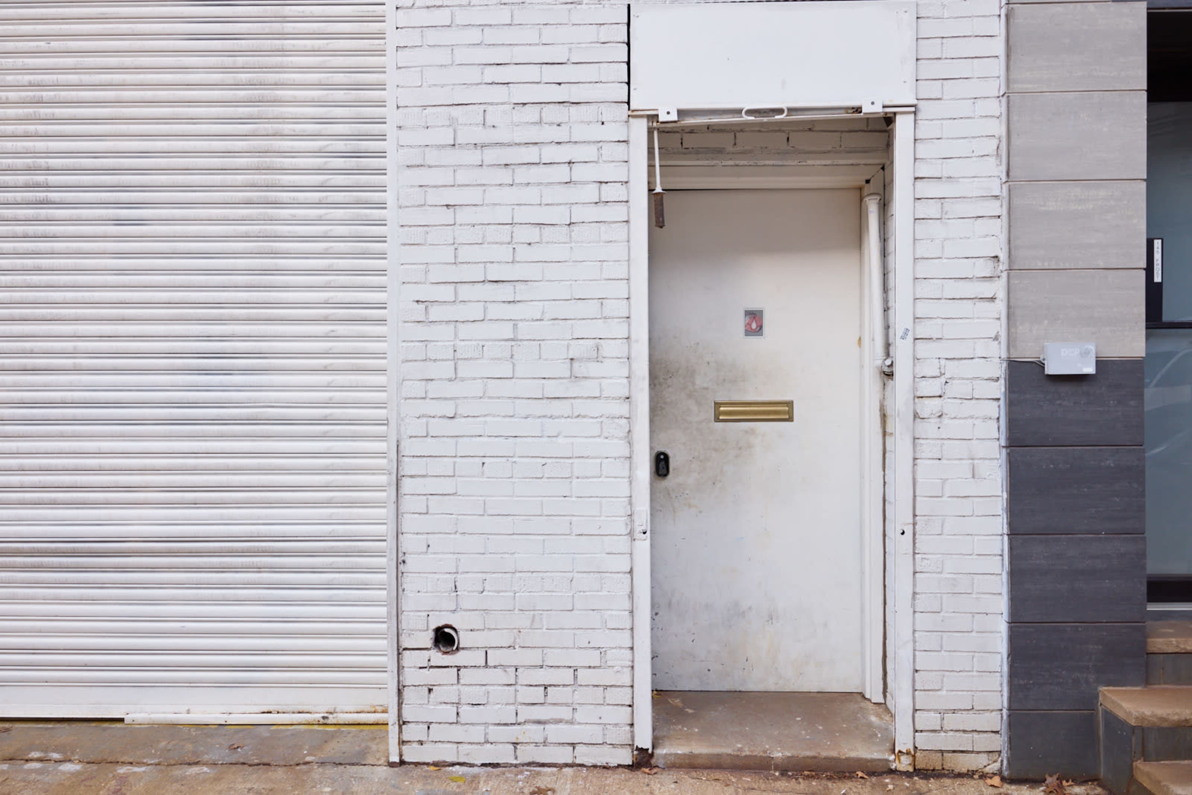 The image shows a white brick wall with a metal roller door to the left and a plain, white door with a mail slot on the right.