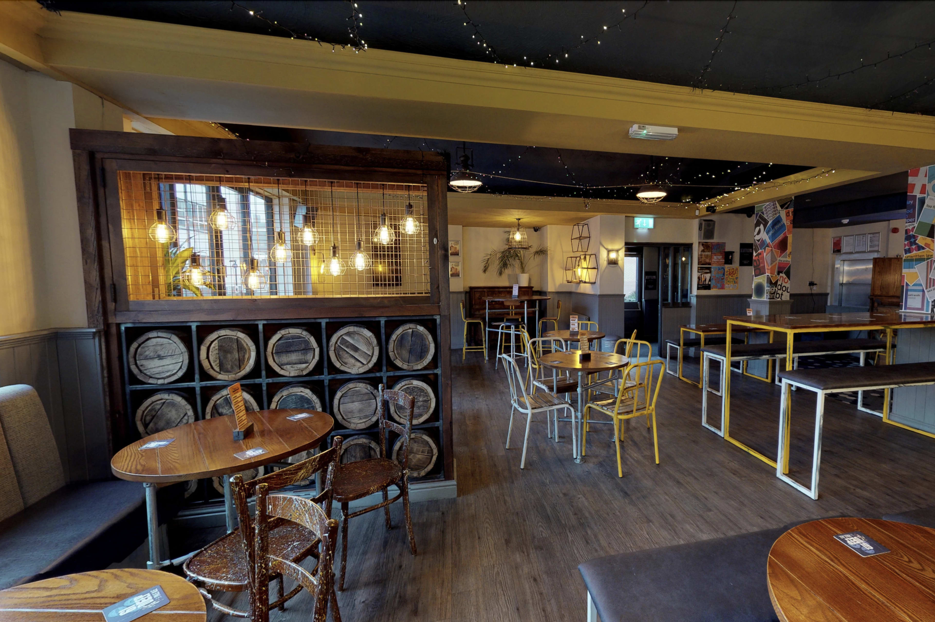 The interior of a pub with wooden flooring, circular tables, and a wall featuring decorative lighting and barrels.