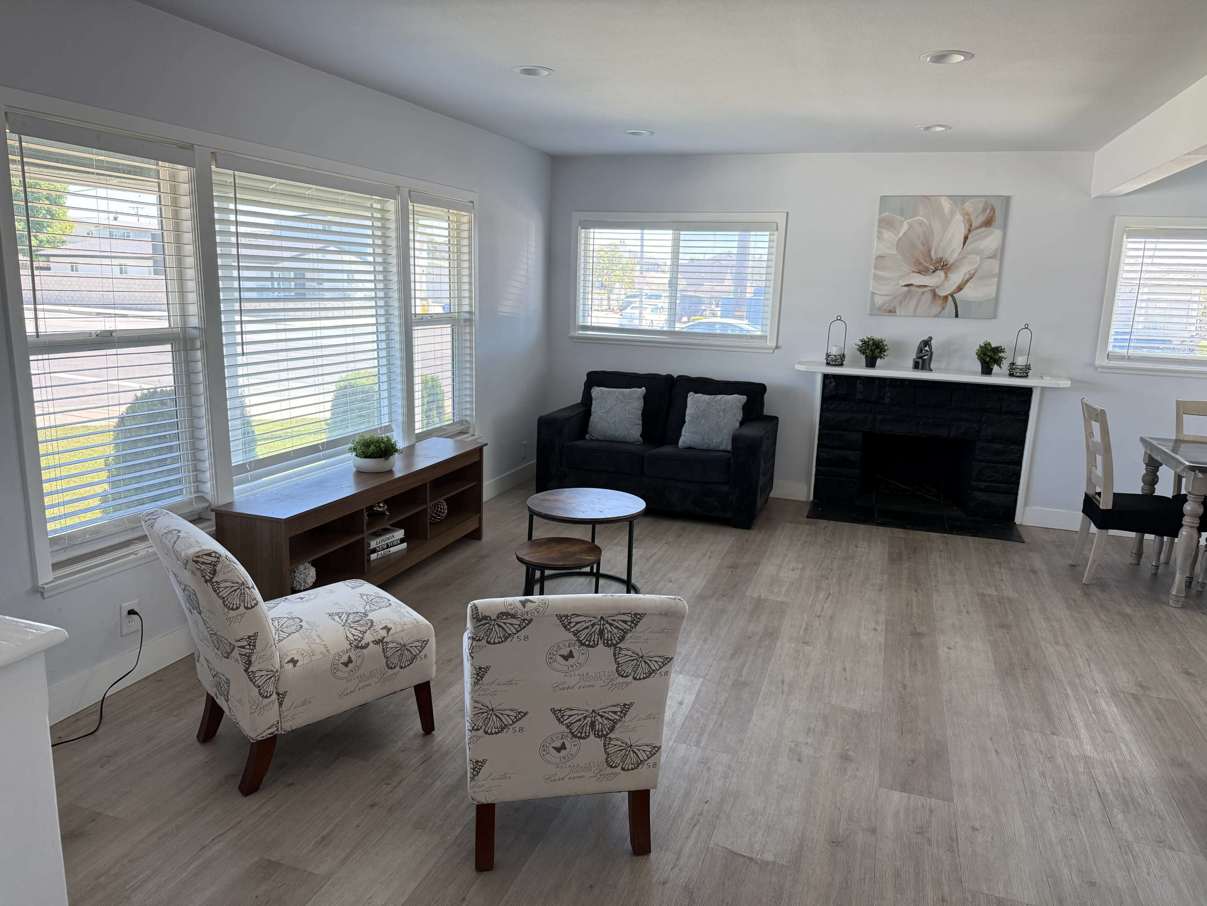 The image shows a bright living room with a seating area featuring two patterned chairs, a small round table, and a black couch, along with a fireplace and dining area visible in the background.