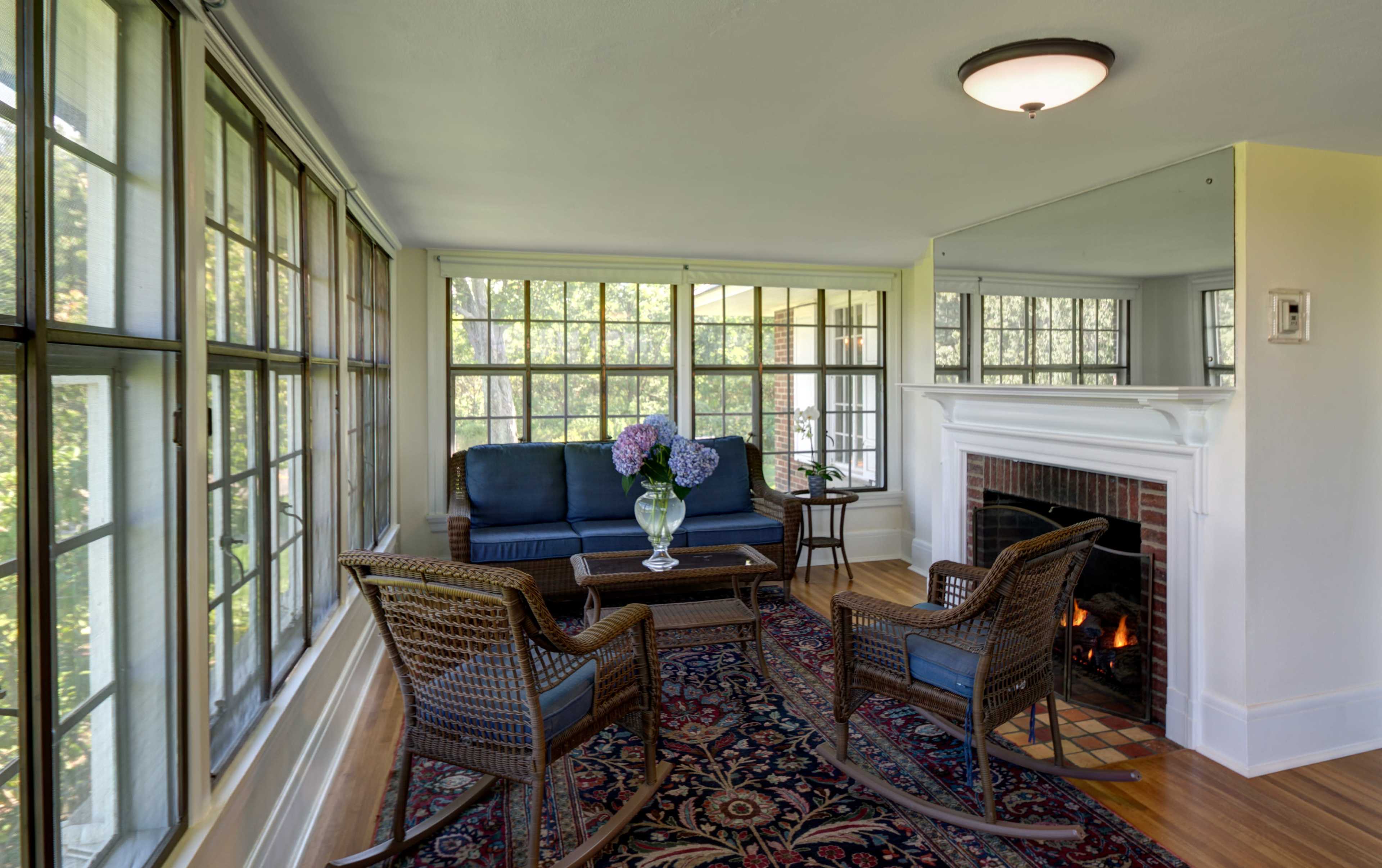 A sunlit living room featuring a blue couch, a coffee table, two wicker chairs, a floral centerpiece, and a fireplace surrounded by large windows.