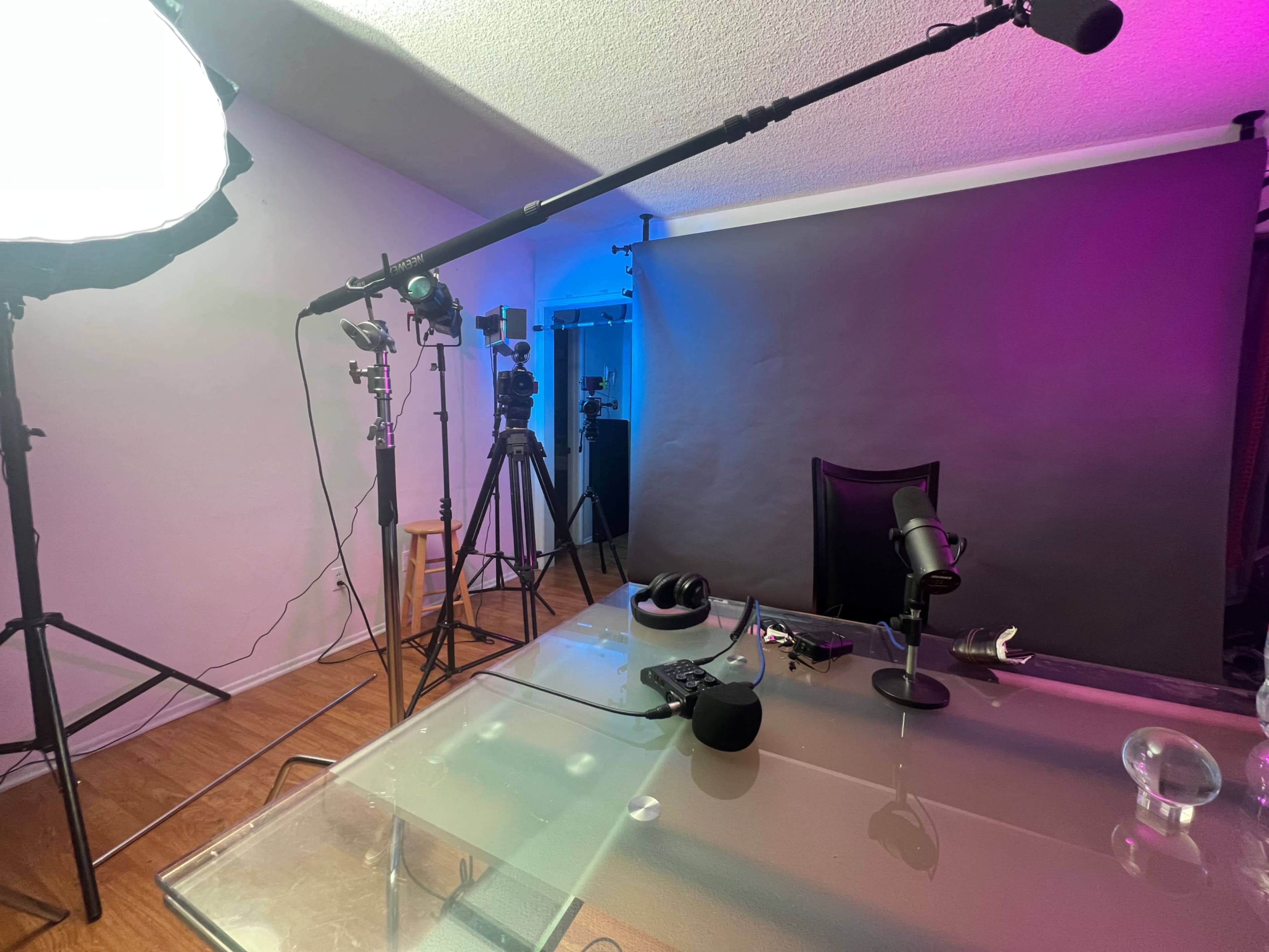 A recording studio setup features a glass table, microphones, cameras on tripods, and colored lighting against a backdrop.