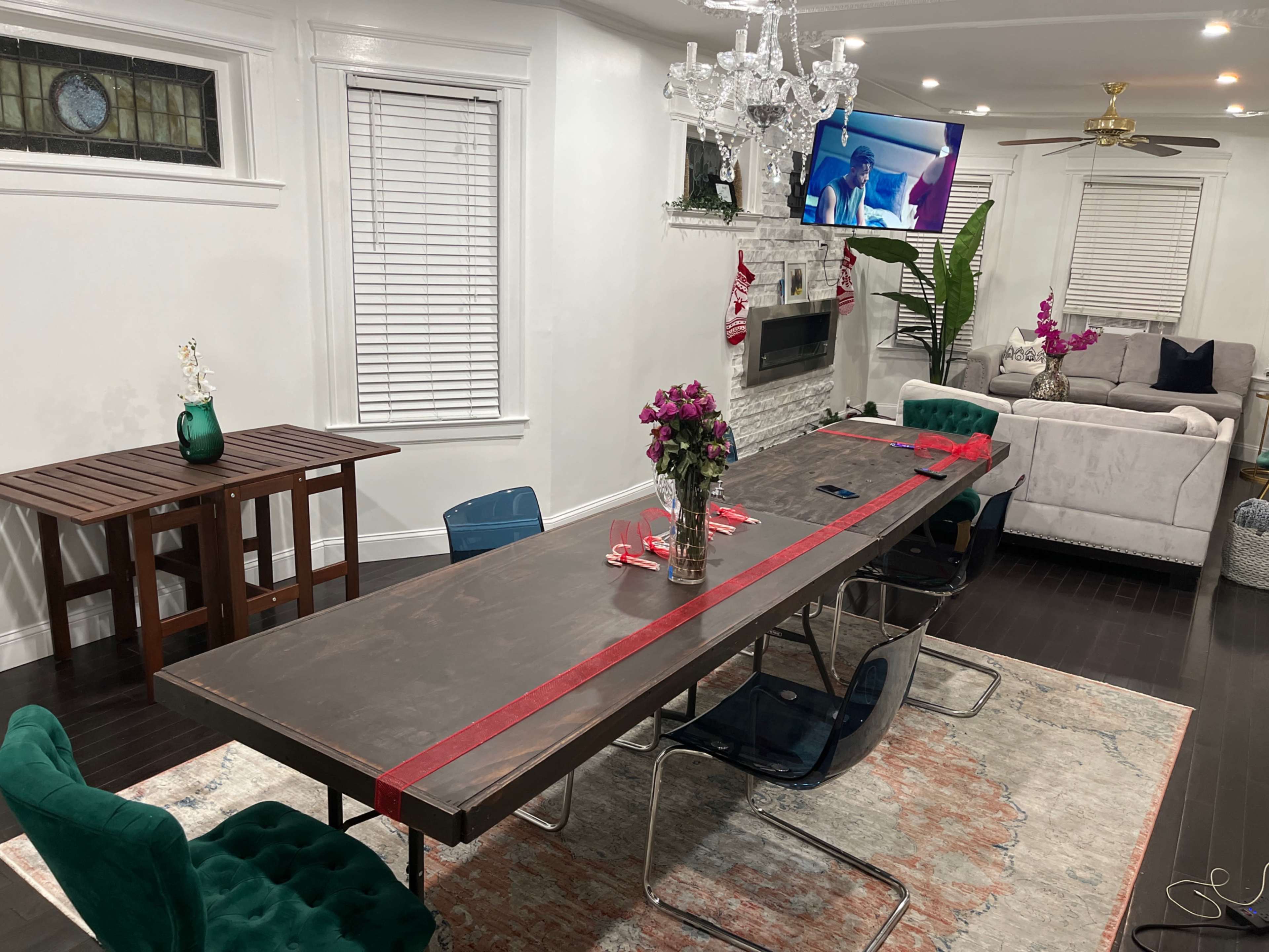 A long dining table is set with red ribbon and flowers, surrounded by chairs, in a room featuring a television, a sofa, and decorative plants.