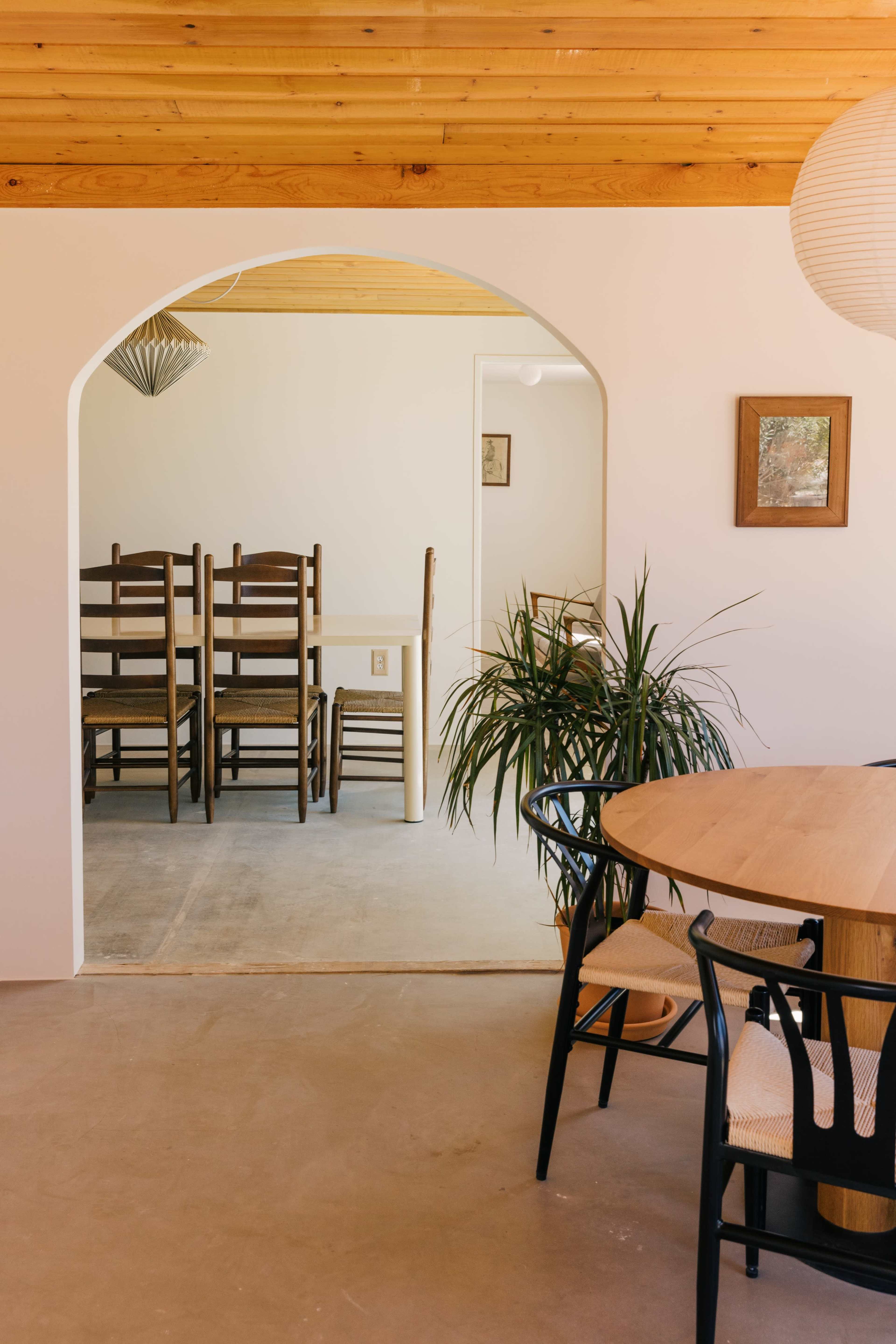 The image shows a spacious interior with a wooden ceiling, a dining area featuring a round table and chairs, and an adjoining room with stacked wooden chairs and a potted plant.