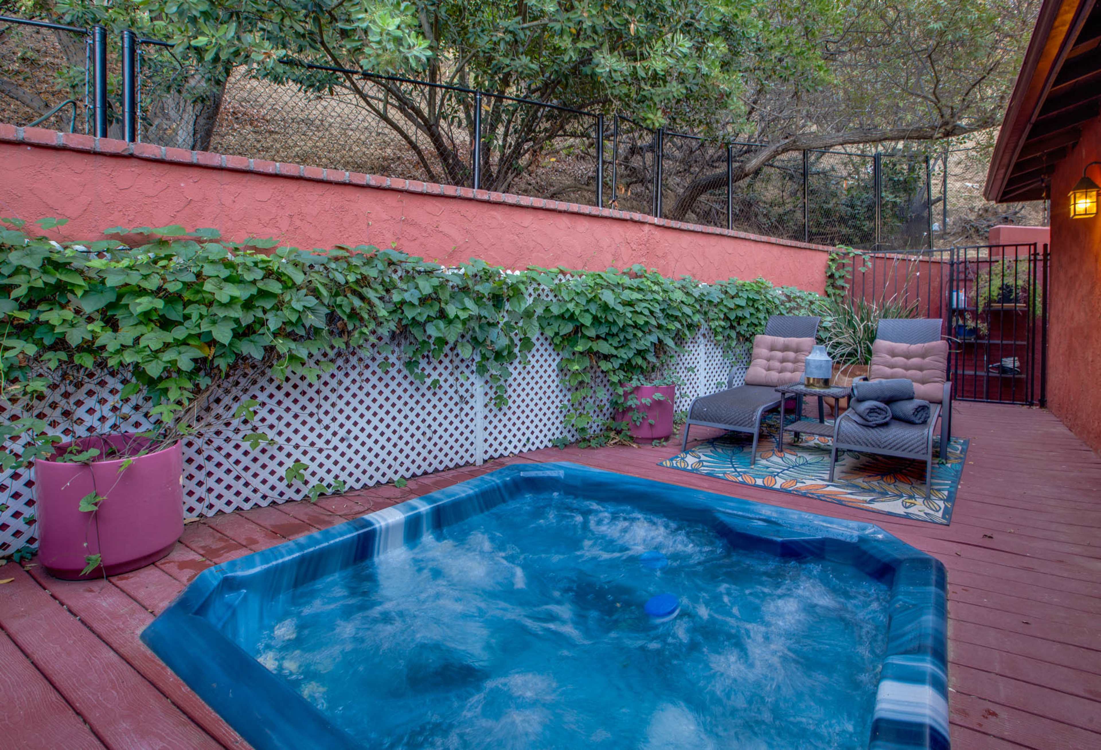 The image shows a small hot tub surrounded by a wooden deck, with lounging chairs and greenery along a red wall.