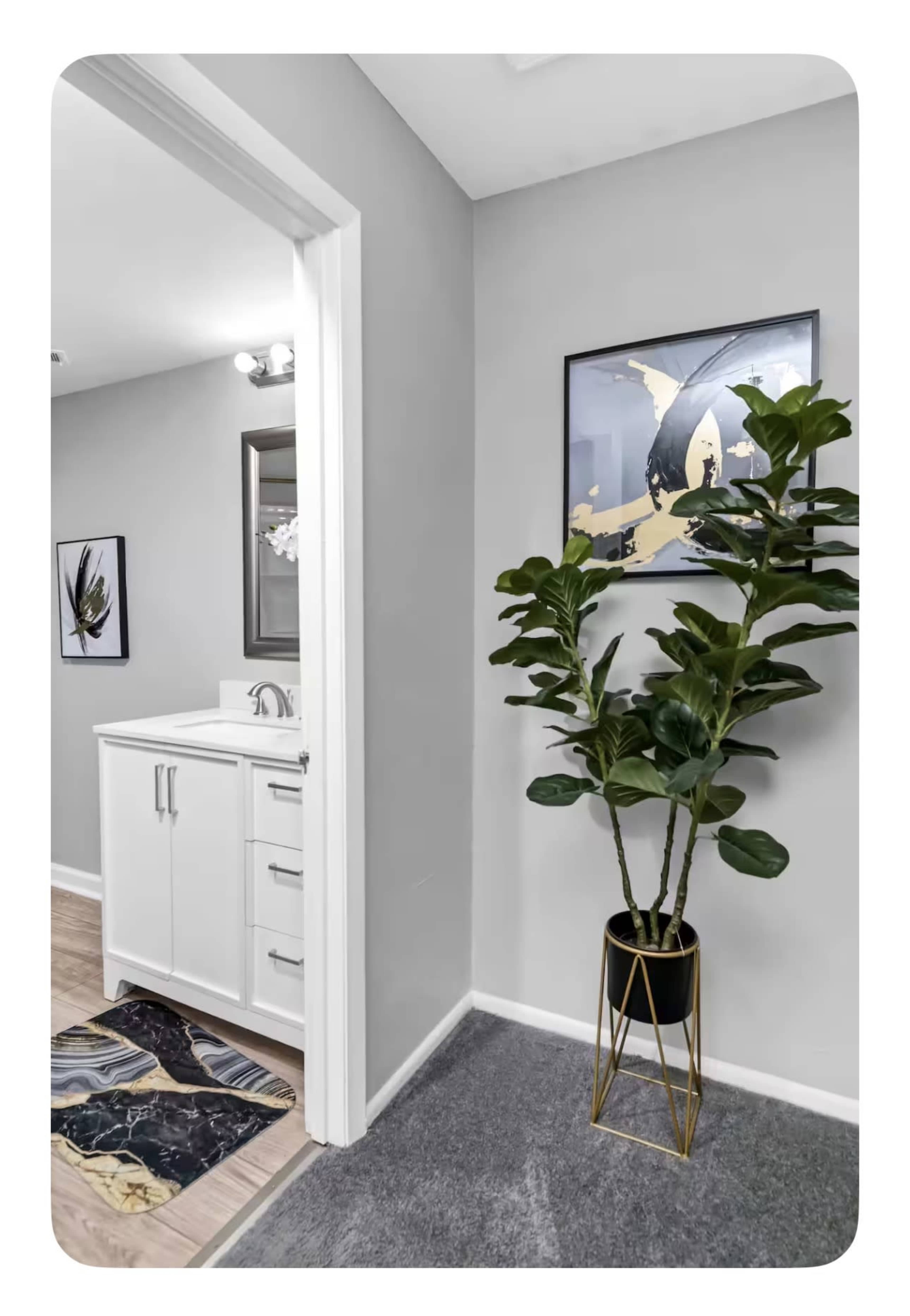 The image shows a bathroom entry with a white vanity, a decorative plant in a black pot, and a framed artwork on the wall.