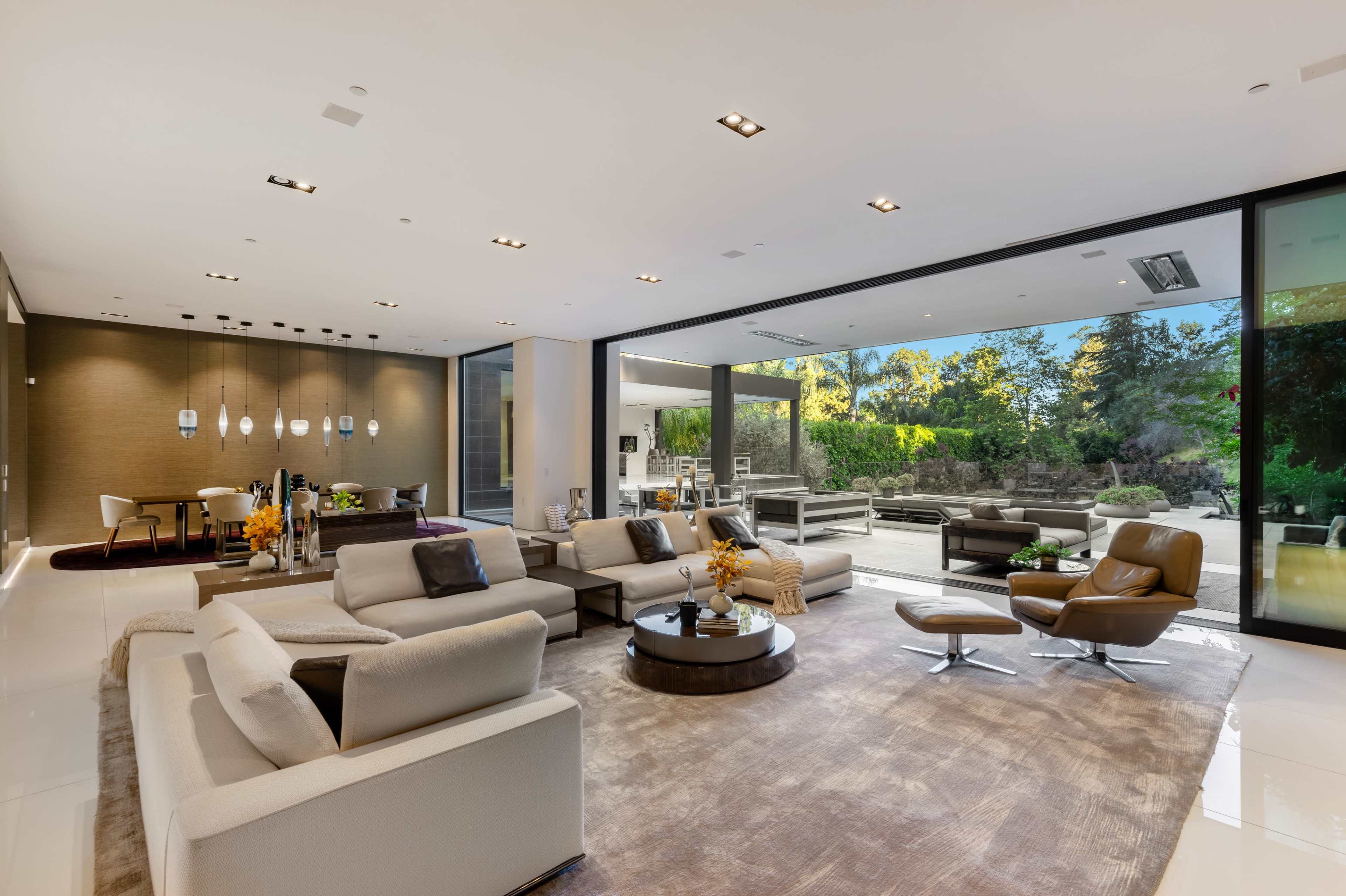 The image shows a modern living room with large glass windows providing a view of an outdoor patio and garden, featuring a beige sectional sofa, a round coffee table, and various contemporary furnishings.