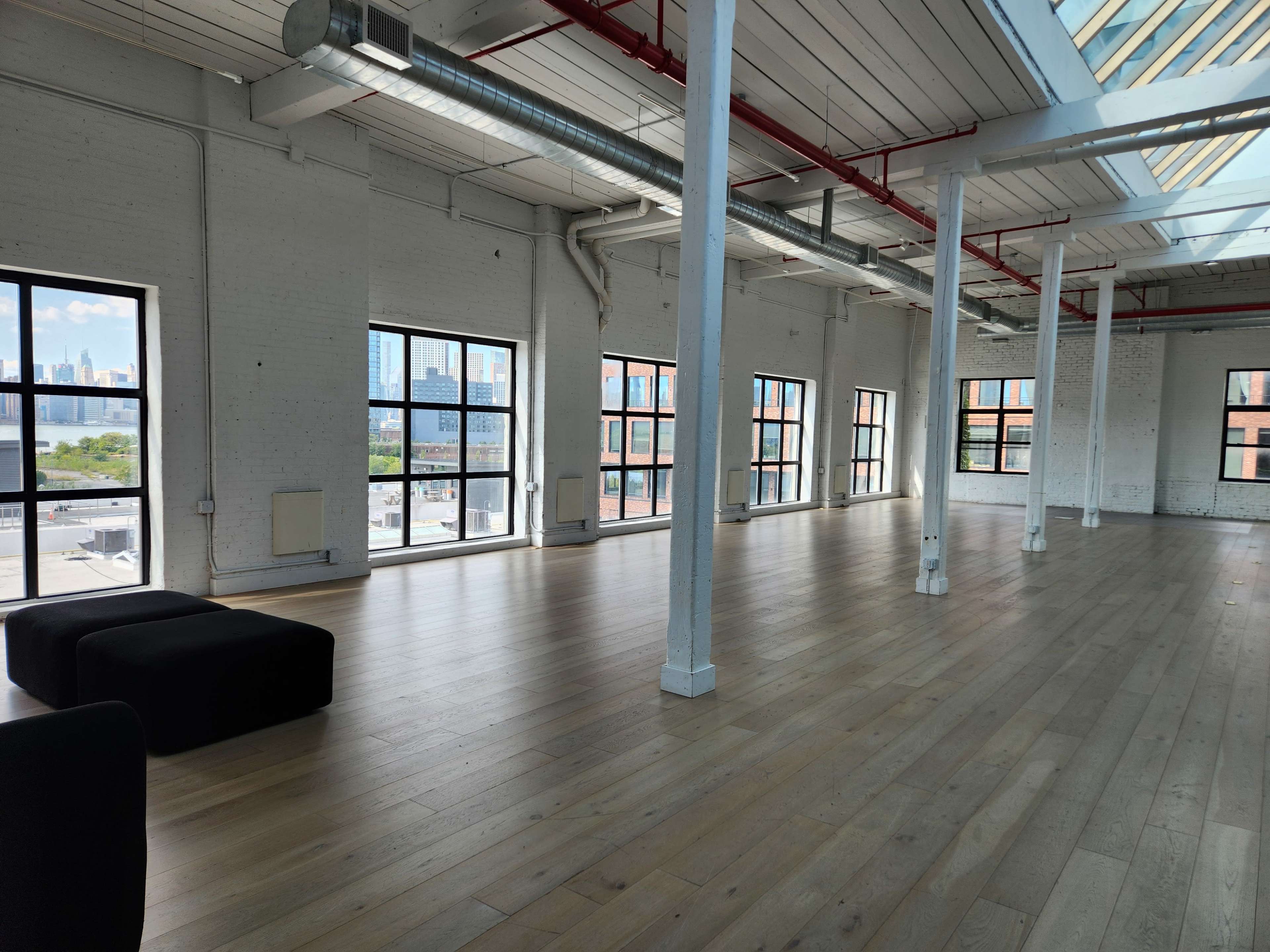 The image shows a spacious, well-lit interior with wooden flooring, large windows, and white walls, featuring two black benches along one side.