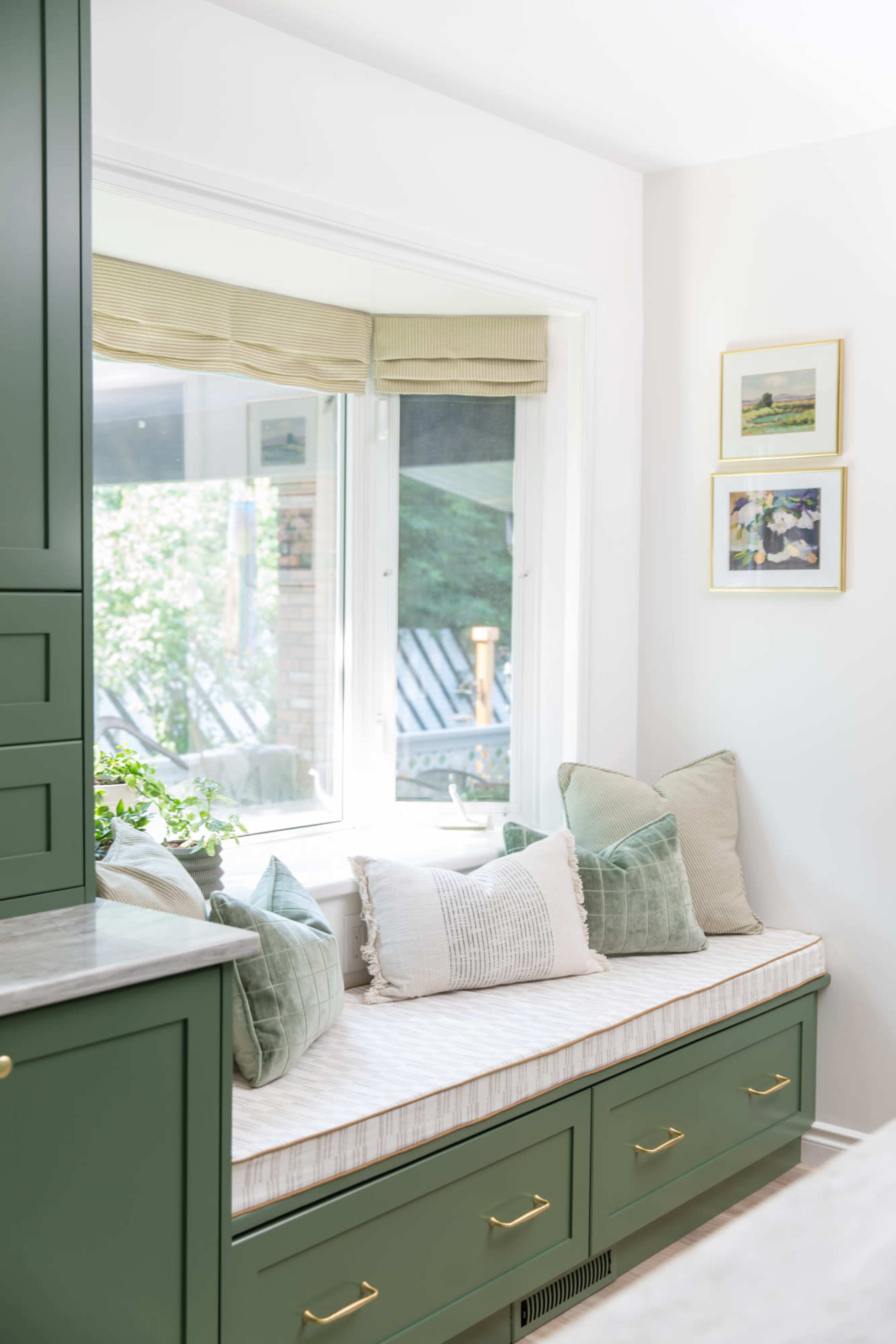 A cozy window seat with decorative pillows is nestled beneath a large window, surrounded by natural light and greenery.