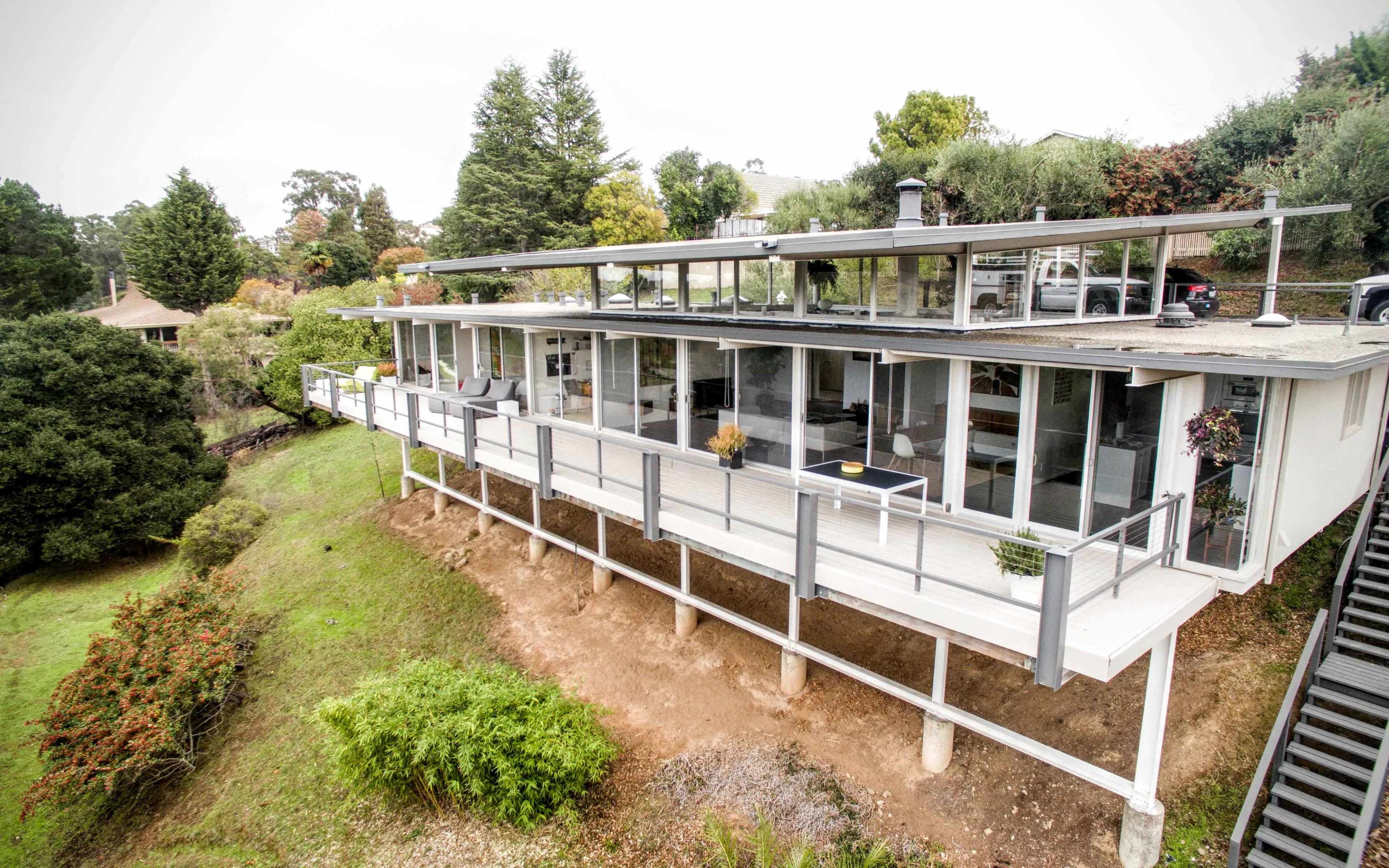 A modern house with large glass windows and a flat roof is positioned on stilts on a hillside, surrounded by greenery.