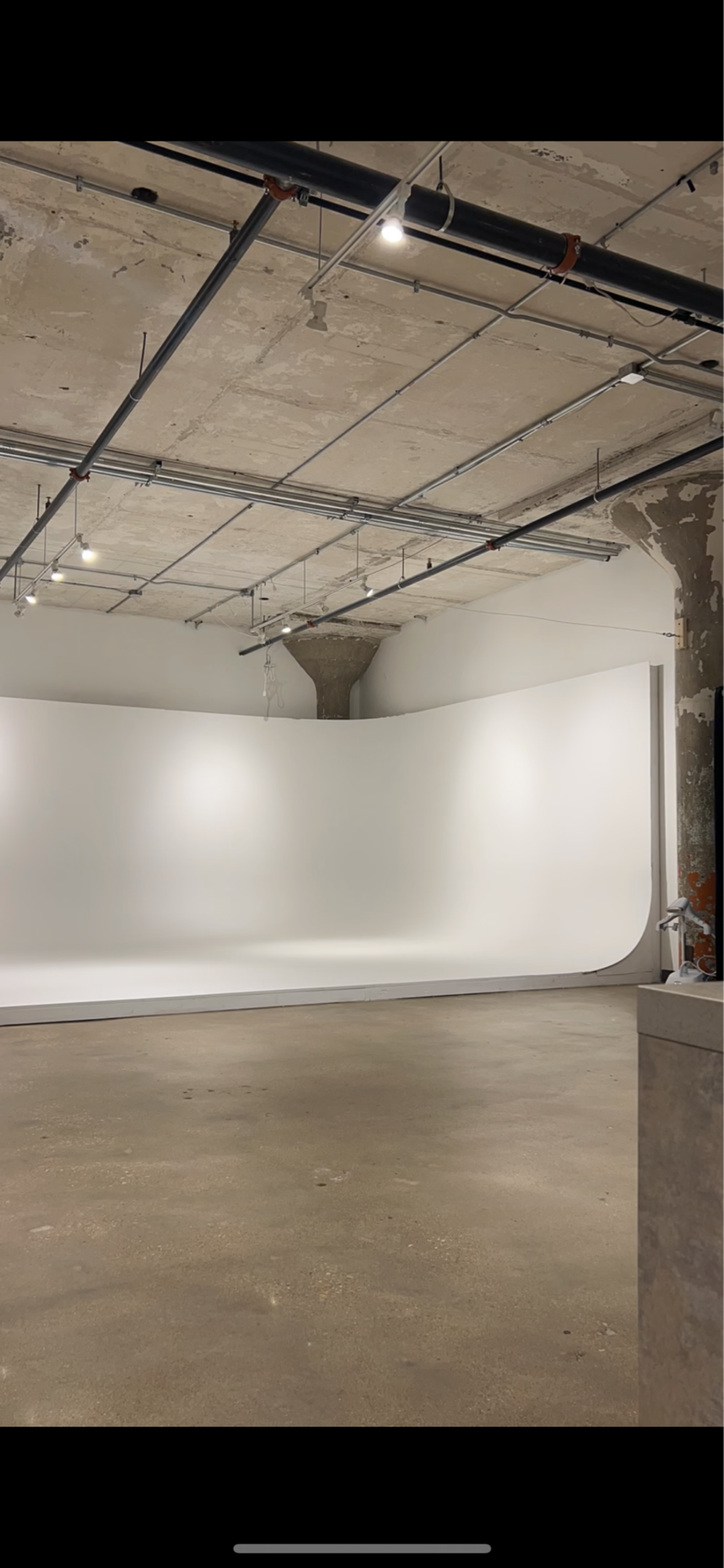The image shows a spacious photography studio with a seamless white backdrop, exposed concrete walls, and overhead lighting.