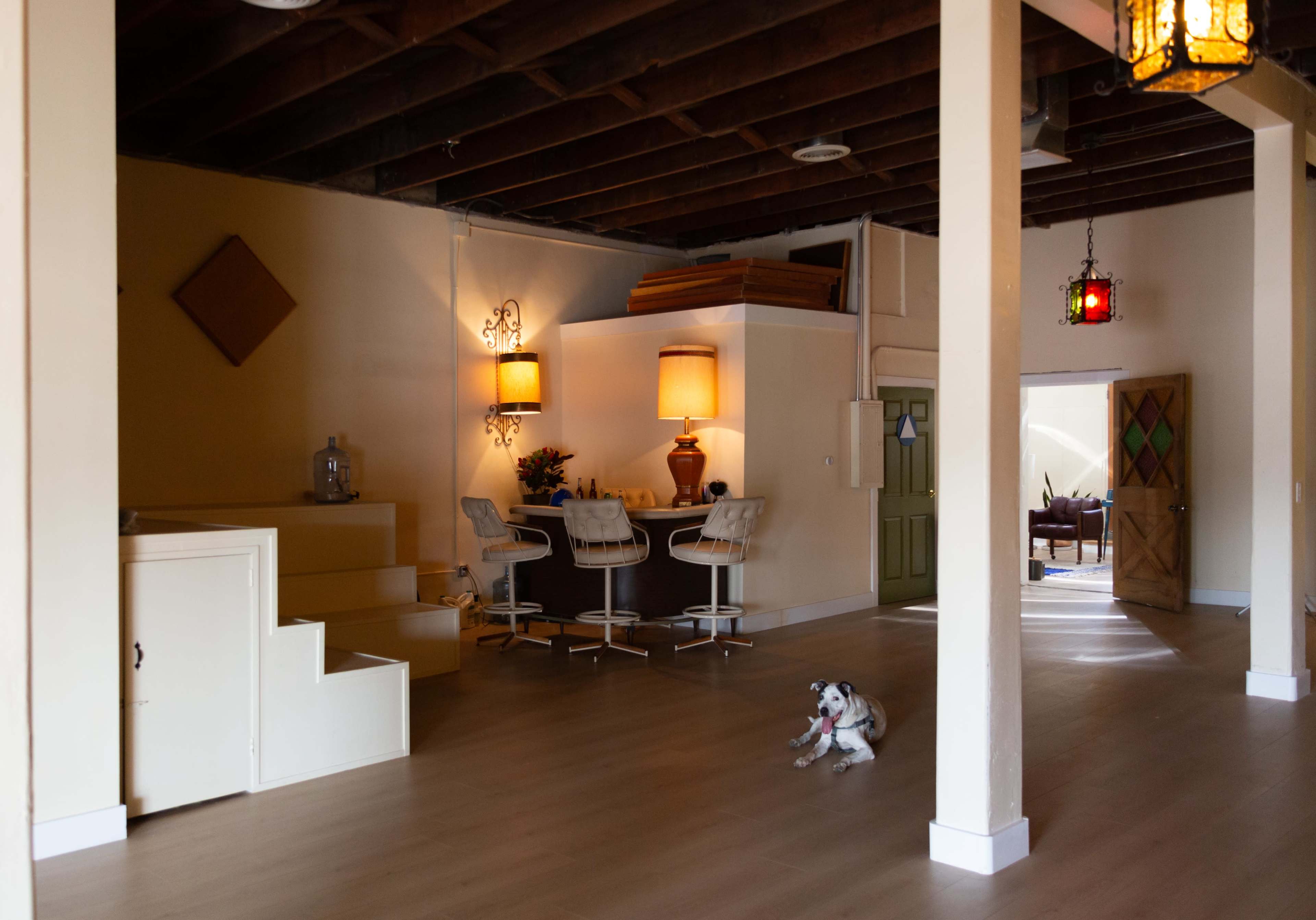 A spacious interior with wooden beams, featuring a seating area with bar stools, a dog on the floor, and an entrance leading to another room.
