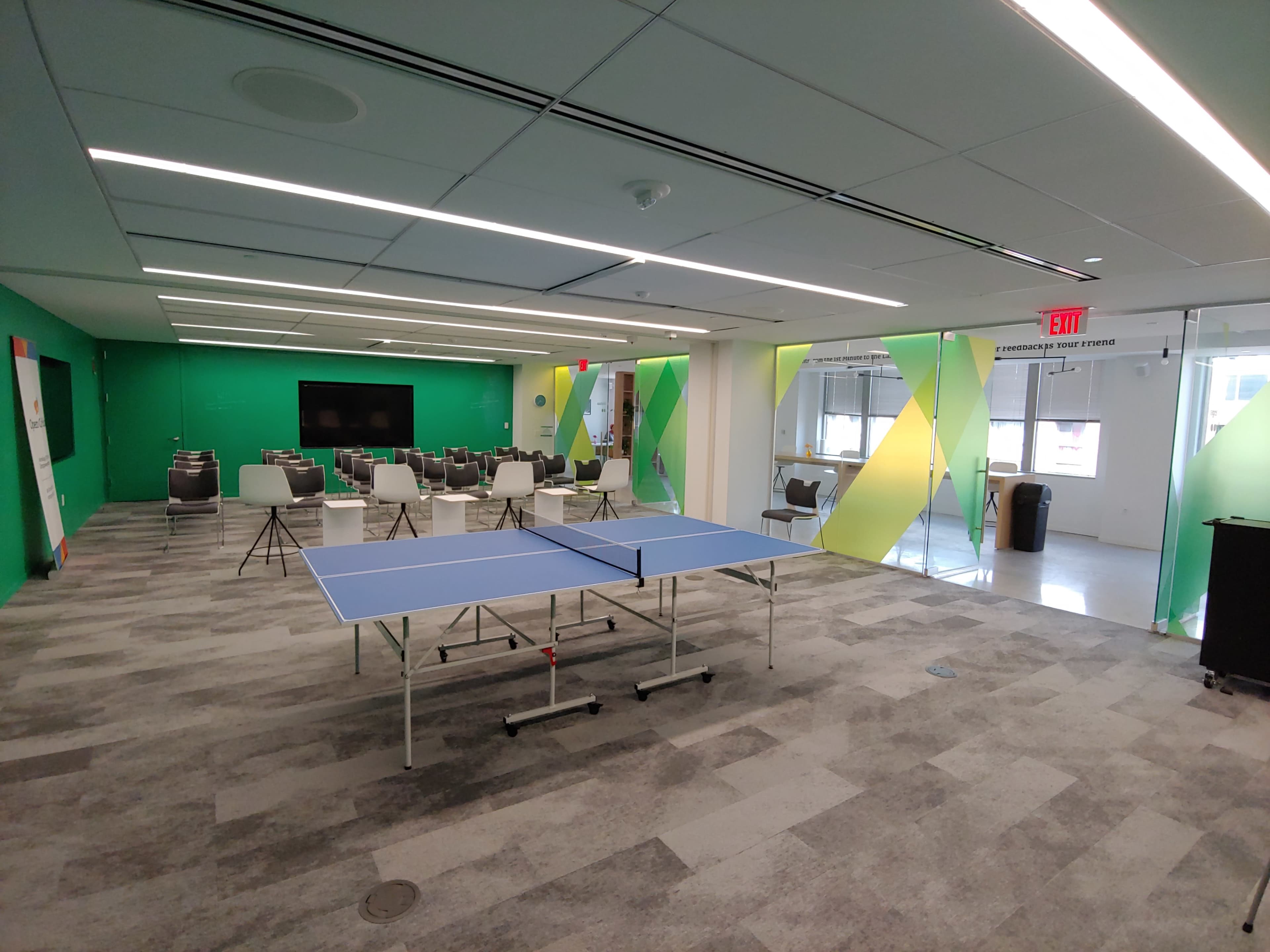 A modern room with a ping pong table in the center, surrounded by chairs and a large screen at one end, with green and yellow walls.