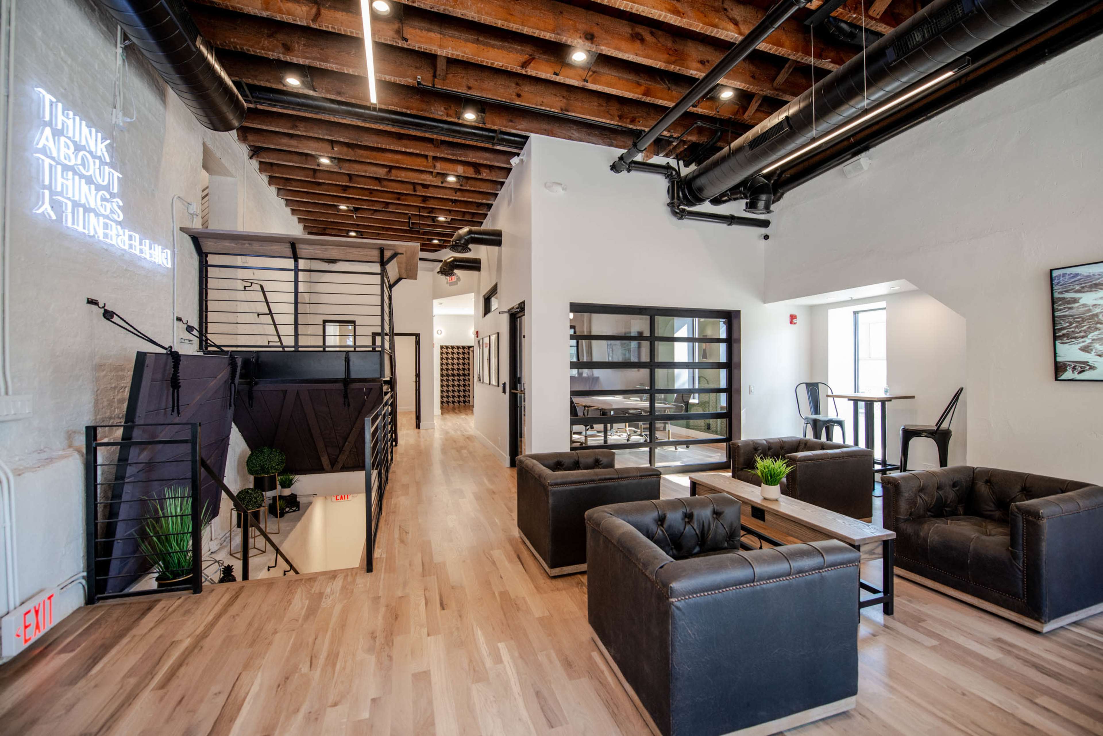The image shows a modern, open office space featuring a lounge area with black sofas, wooden floors, exposed ceiling beams, and a sleek staircase.