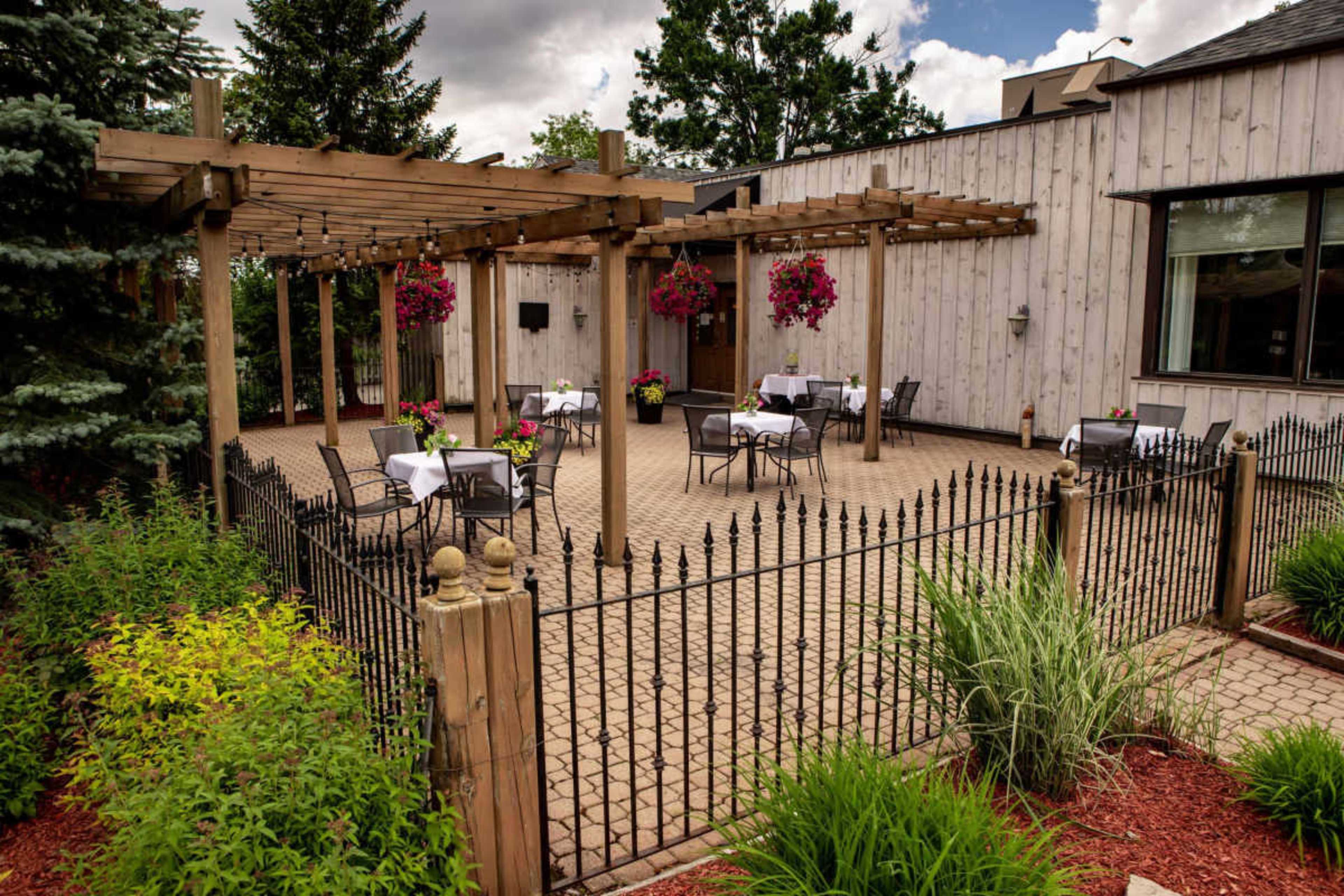 The image shows an outdoor dining area with several tables and chairs arranged under a wooden pergola, surrounded by landscaped greenery and flower planters.