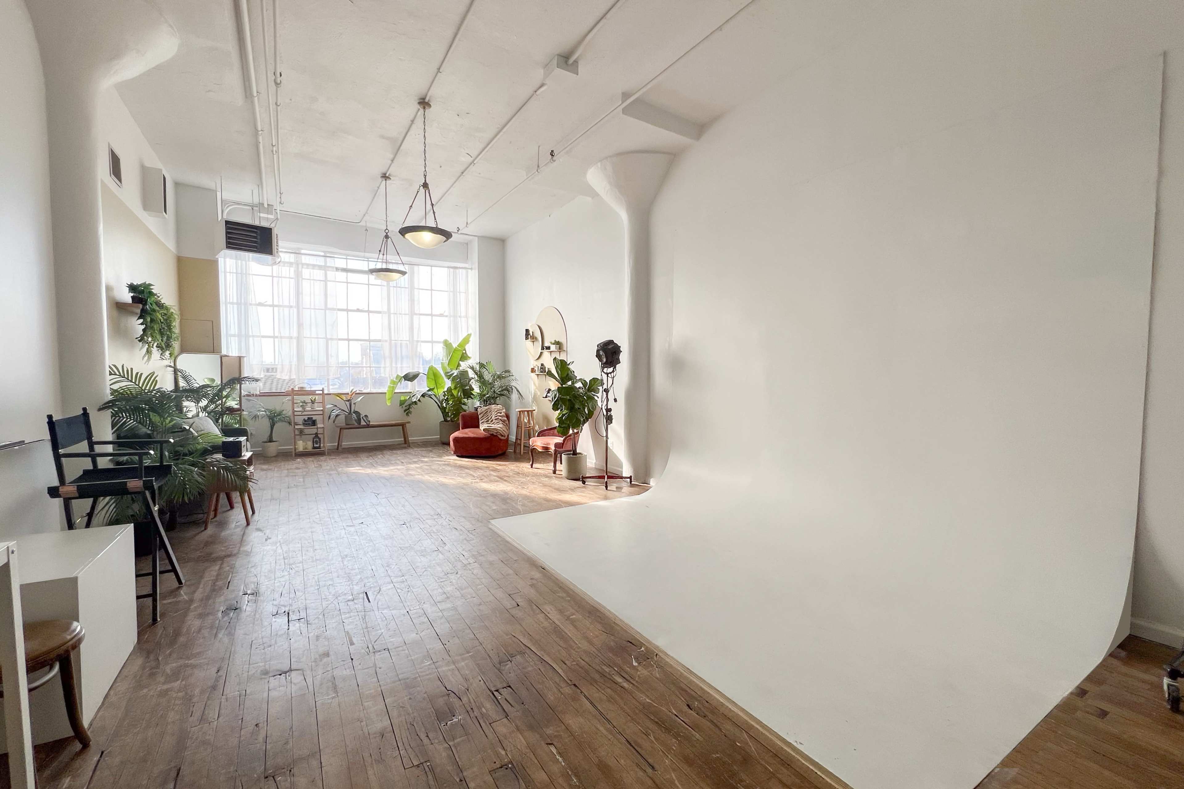 The image shows a spacious studio with a white backdrop, wooden flooring, and various plants, furniture, and natural light coming through large windows.