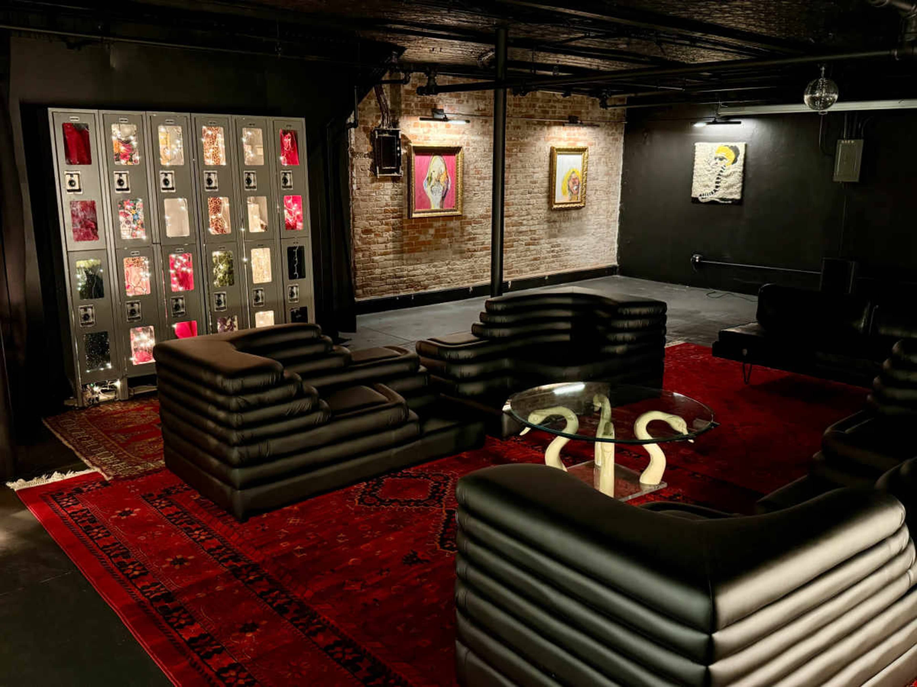 The image shows a modern lounge area featuring sleek black furniture, a glass coffee table, and artwork displayed on the brick walls, all set against a dark floor and illuminated by overhead lights.