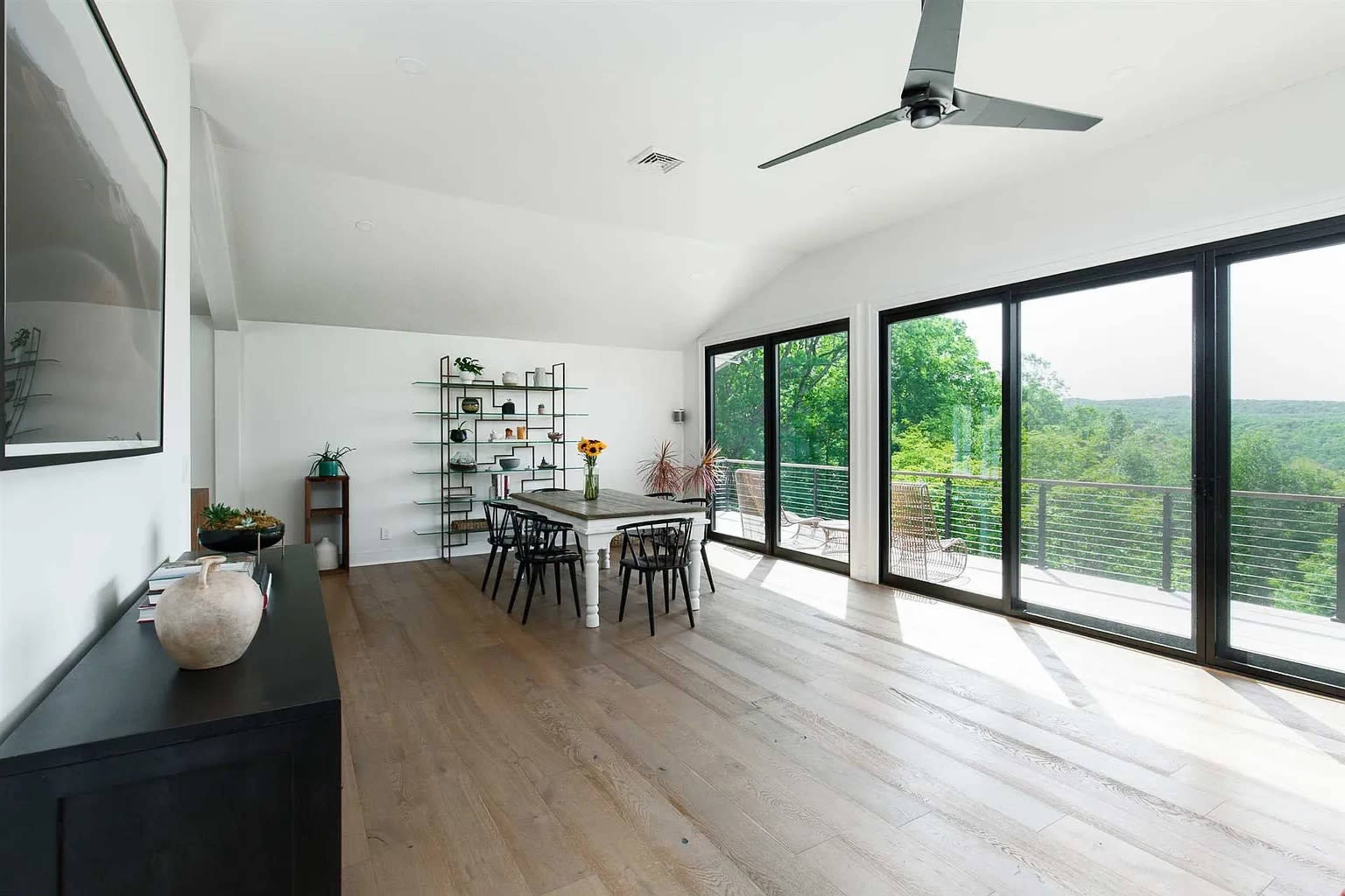 A modern dining area features a long table surrounded by black chairs, large windows offering a view of greenery, and light wood flooring.