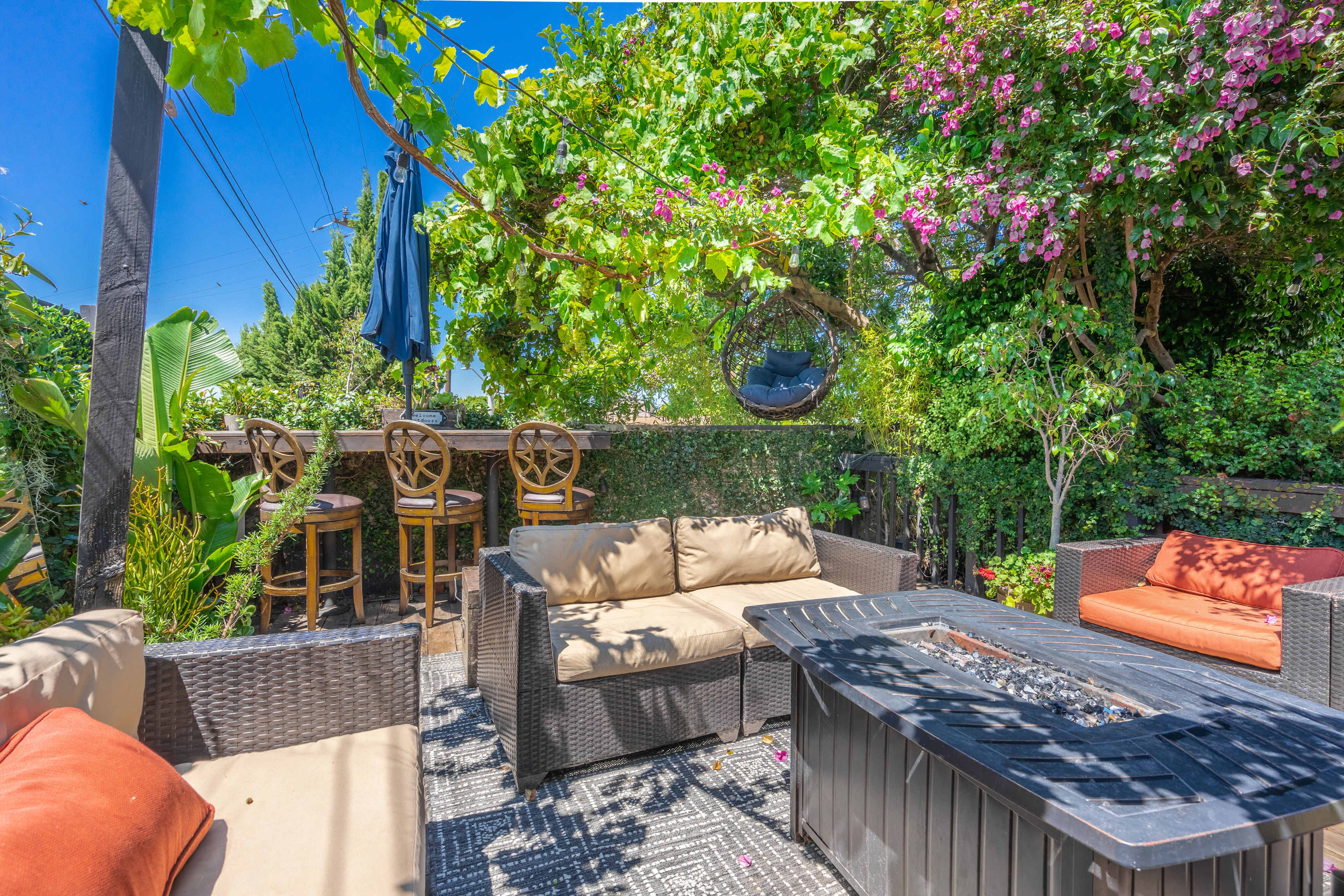 A cozy outdoor living space features a fire pit surrounded by comfortable seating and a bar area with high stools, all set against a backdrop of greenery and flowering plants.