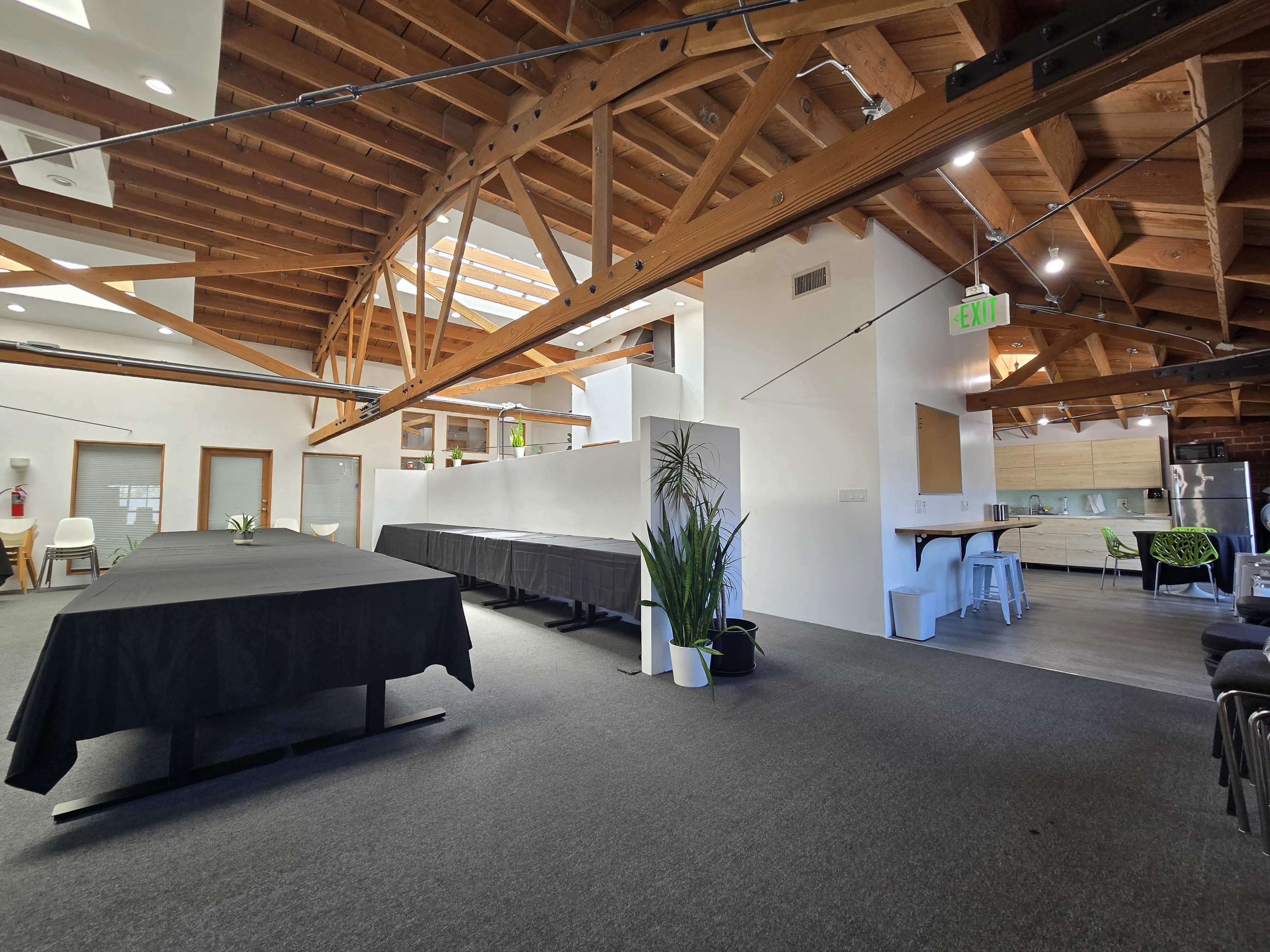 The image shows a spacious office interior with exposed wooden beams, a long black table in the foreground, and various seating areas in a modern setting.