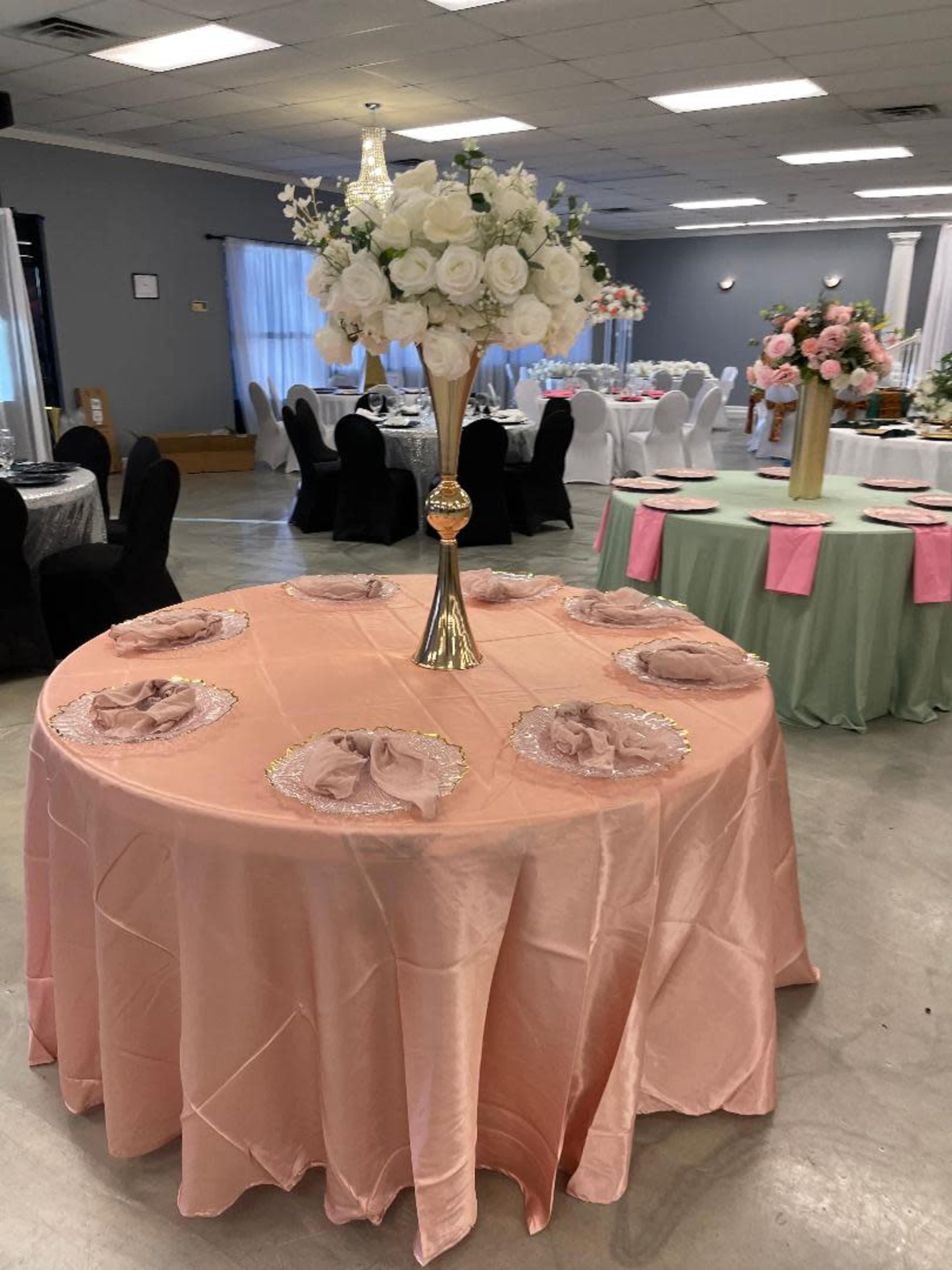 A round table covered in a pink tablecloth is set with decorative plates and napkins, topped with a tall floral centerpiece.