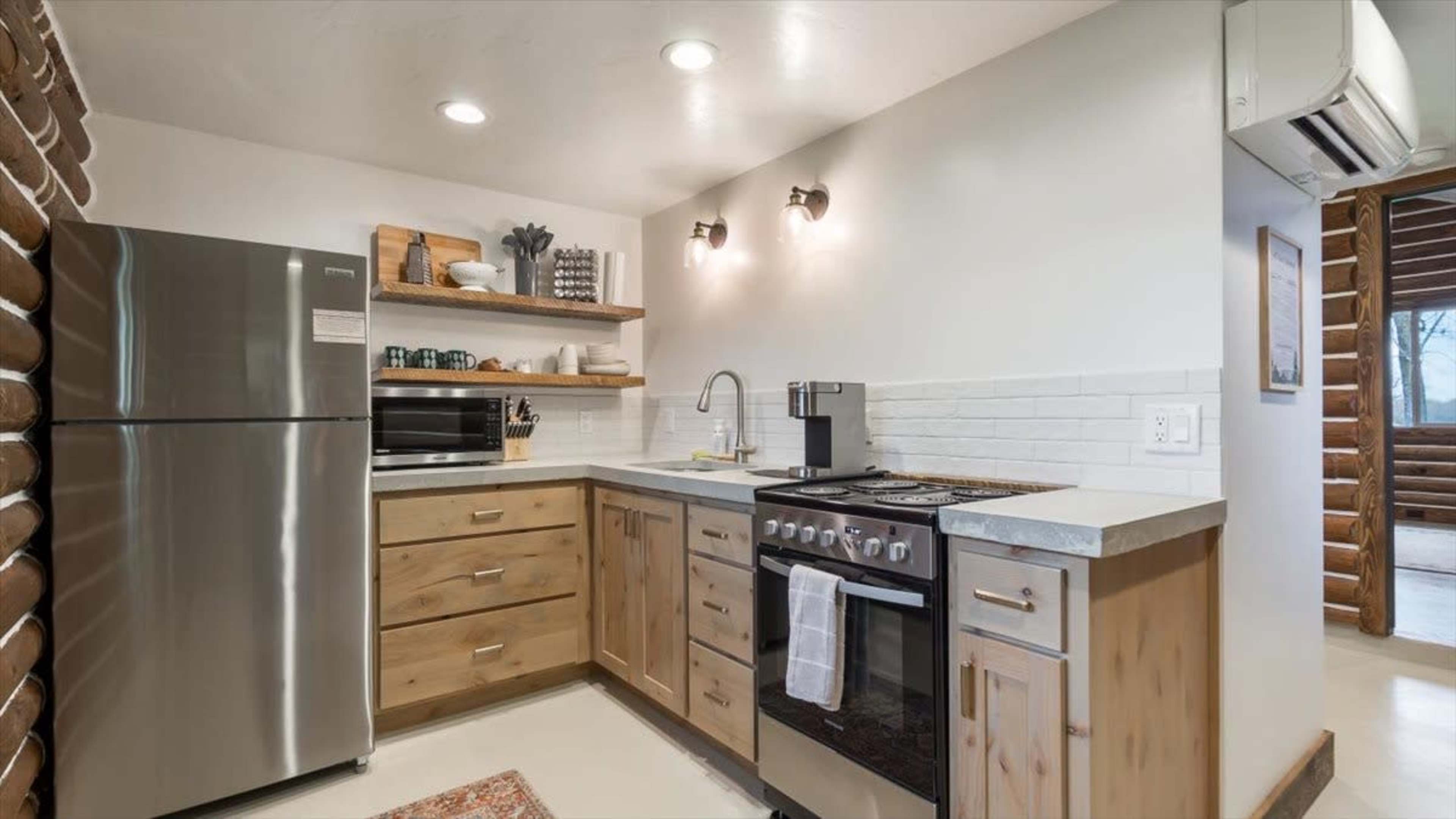 The image shows a modern kitchen with wooden cabinets, a stainless steel refrigerator, a stove, a microwave, and wall-mounted lights.