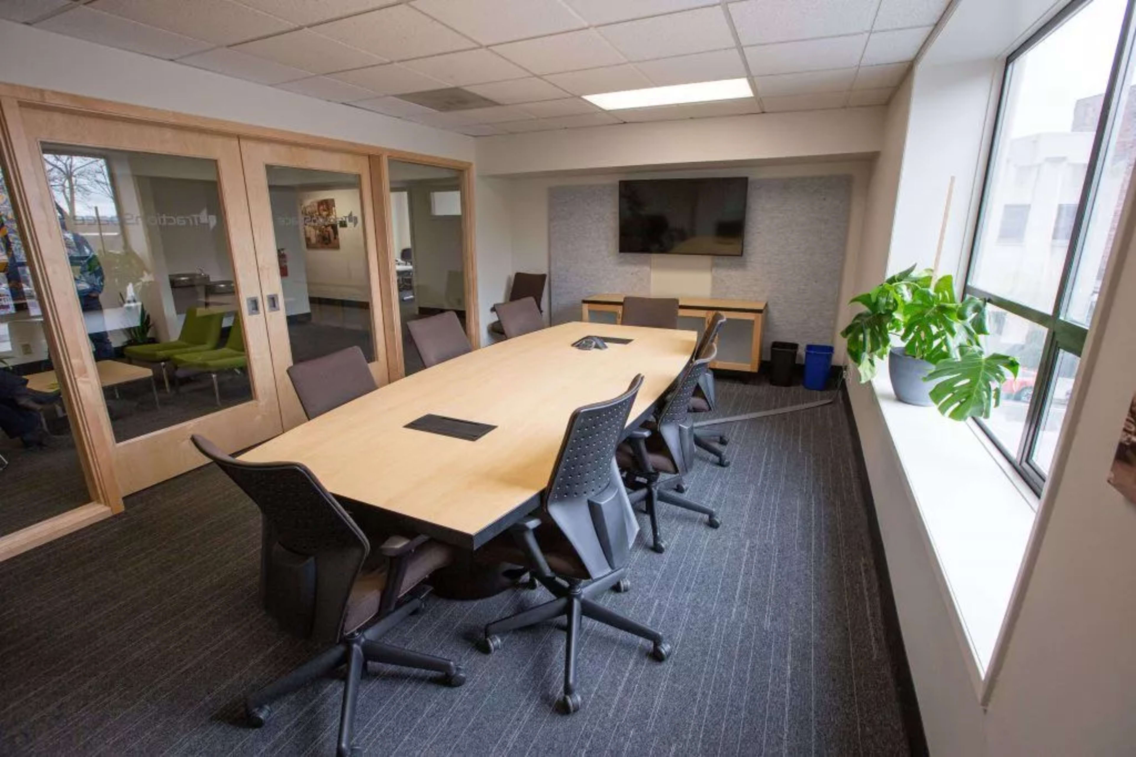 A large conference room features a central wooden table surrounded by office chairs, with a television mounted on the wall and a potted plant near the window.